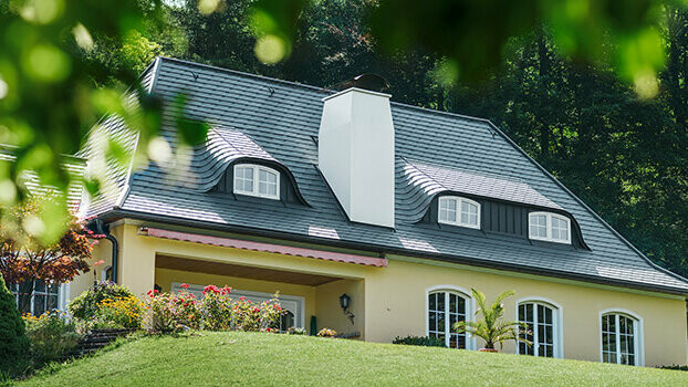 Maison individuelle avec toiture récemment rénovée à l’aide de bardeaux de toiture PREFA couleur anthracite, avec lucarnes arrondies (lucarne à joues galbées) et cheminée blanche.