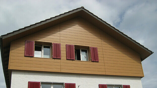 Habillage du pignon d’une maison classique avec toit à deux pans. Le pignon est habillé à l’aide de Sidings PREFA couleur chêne naturel posés à l’horizontale. Les fenêtres disposent de volets de couleur rouge.