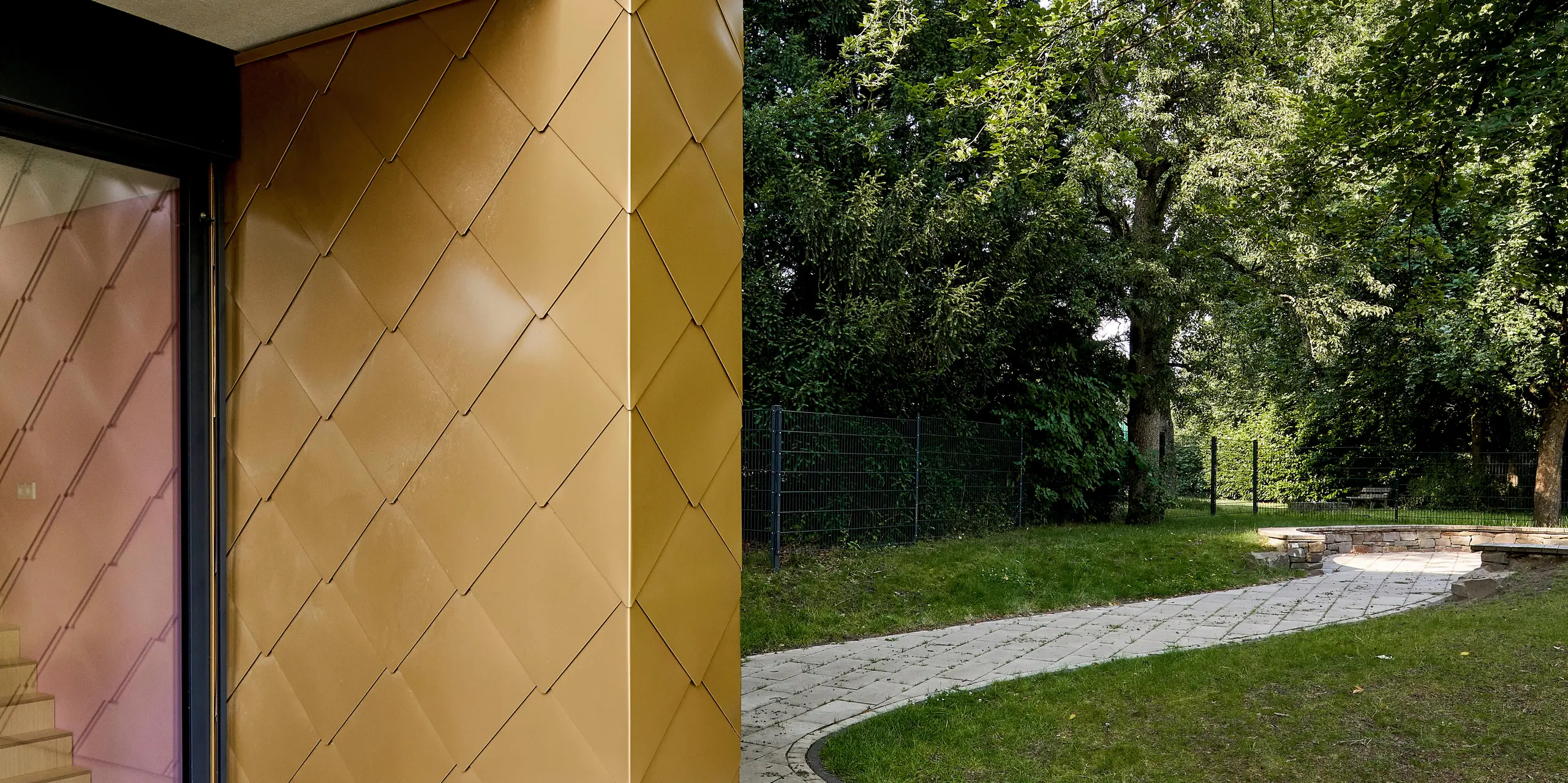 Une architecture moderne, sublimée par une façade en aluminium PREFA or aux reflets dorés et au motif d'écailles, caractérise ce paysage dynamique. Les losange, agencés avec précision, reflètent magnifiquement la lumière du soleil et créent un contraste saisissant avec la végétation environnante. L'élégant balcon aux lignes épurées complète l'ensemble avec raffinement. Durables et résistants aux intempéries, les panneaux muraux PREFA séduisent par leur éclat, leur robustesse et leur conception sans entretien – idéaux pour une conception de façade sophistiquée et durable, aussi bien en milieu urbain que naturel.