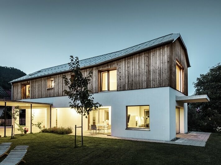 Vue de face de cette maison familiale individuelle à Pruggern, au crépuscule. L'intérieur de la maison est illuminée. La toiture a été revêtue avec le PREFALZ dans la teinte aluminium naturel.