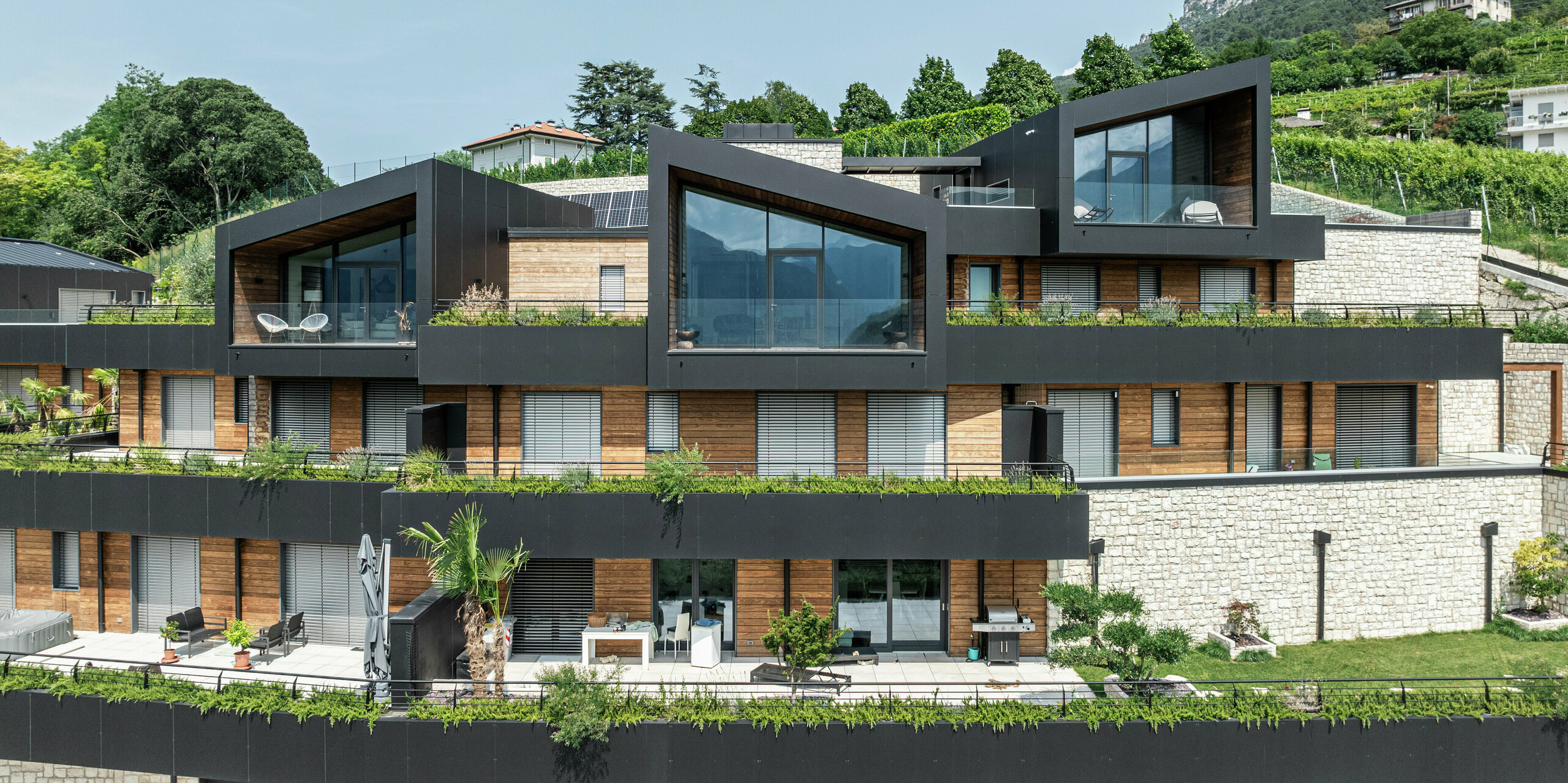 Élégance et fonctionnalité à la perfection : L'architecture saisissante de ce complexe résidentiel se caractérise par des pignons modernes, de grandes surfaces vitrées et un mélange harmonieux de matériaux. La façade en panneaux composites en aluminium PREFA en P.10 Noir assure une enveloppe de bâtiment durable et sans entretien, tandis que le toit PREFALZ offre une protection optimale. Le bois et la pierre naturelle ajoutent des accents chaleureux et se fondent parfaitement dans l'environnement verdoyant. Des terrasses verdoyantes et des espaces de vie spacieux créent une expérience de vie unique avec un confort maximal et des vues panoramiques.