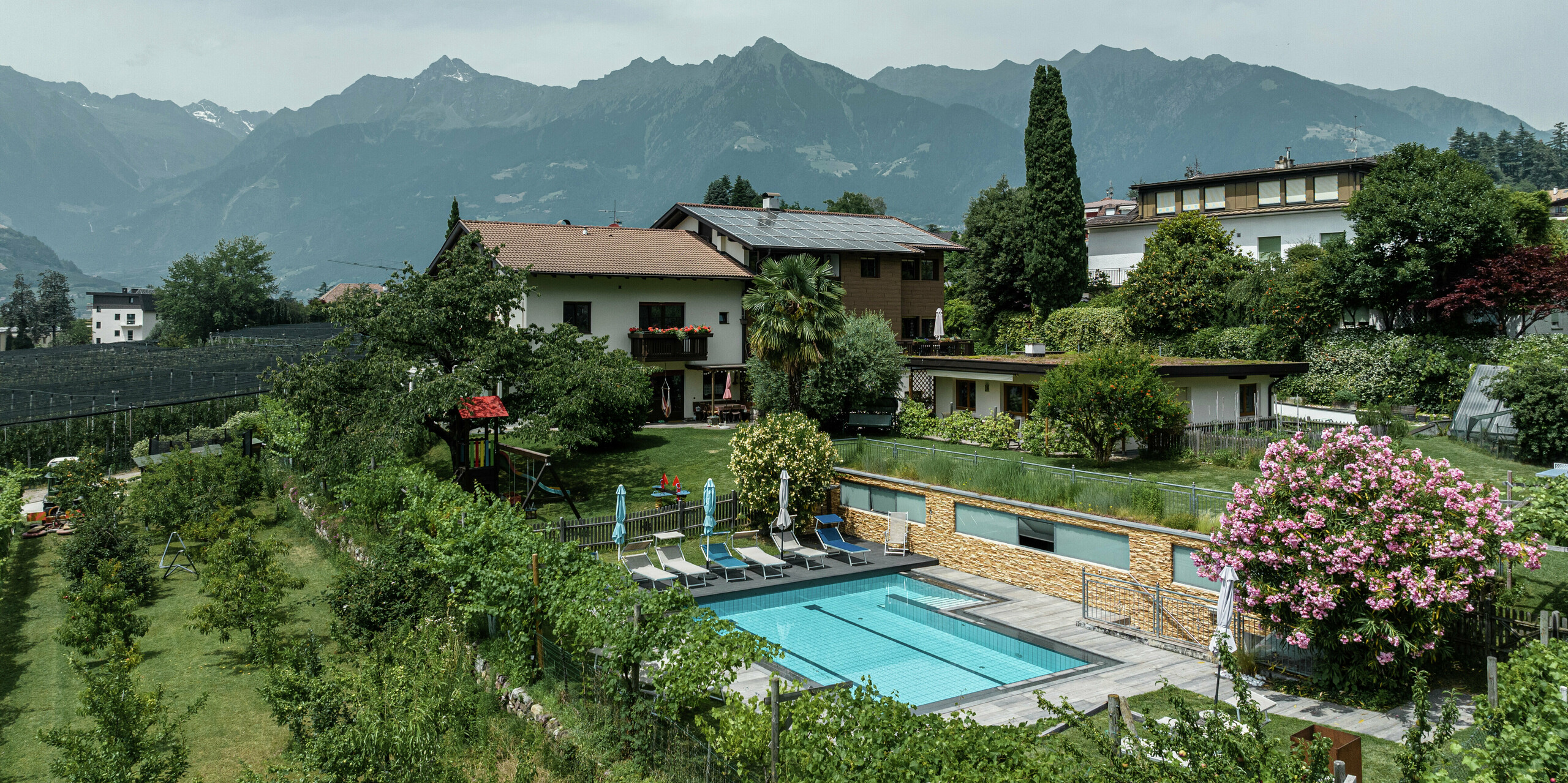 Appartements de vacances Prechtlhof à Merano, dans le Tyrol du Sud, entourés de vergers et de panoramas de montagnes. La façade rénovée avec des Sidings PREFA en noyer foncé s'intègre harmonieusement dans le paysage. Au premier plan, un espace piscine avec chaises longues et fleurs colorées vous invite à la détente.