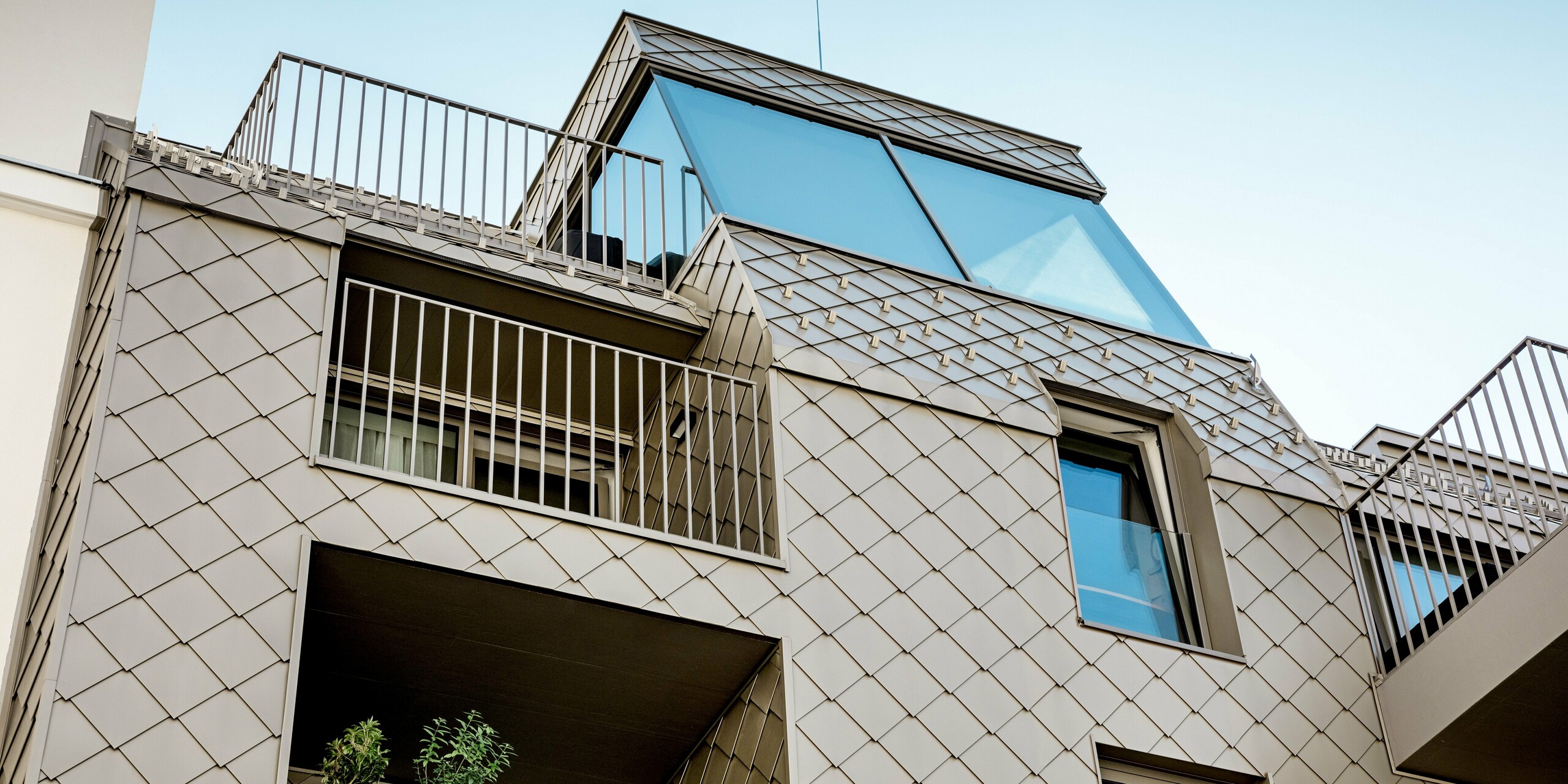 Das Bild zeigt die beeindruckende Erweiterung eines Wohngebäudes in der Rötzergasse in Wien, bei der PREFA Rauten 29×29 in Bronze für das Dach und die Innenhoffassade verwendet wurden. Die modernen Aluminiumrauten bilden eine harmonische Verbindung zur traditionellen Architektur der Nachbargebäude und setzen dabei spannende Akzente. Besonders auffällig sind die präzise verlegten Rauten, die an den Dachkanten und über die Loggien hinweg laufen, und dabei sowohl funktionale als auch ästhetische Anforderungen perfekt erfüllen. Die bronzefarbene Oberfläche der Rauten harmoniert ideal mit den Fensterprofilen und Geländern und sorgt für eine stilvolle Gesamterscheinung.