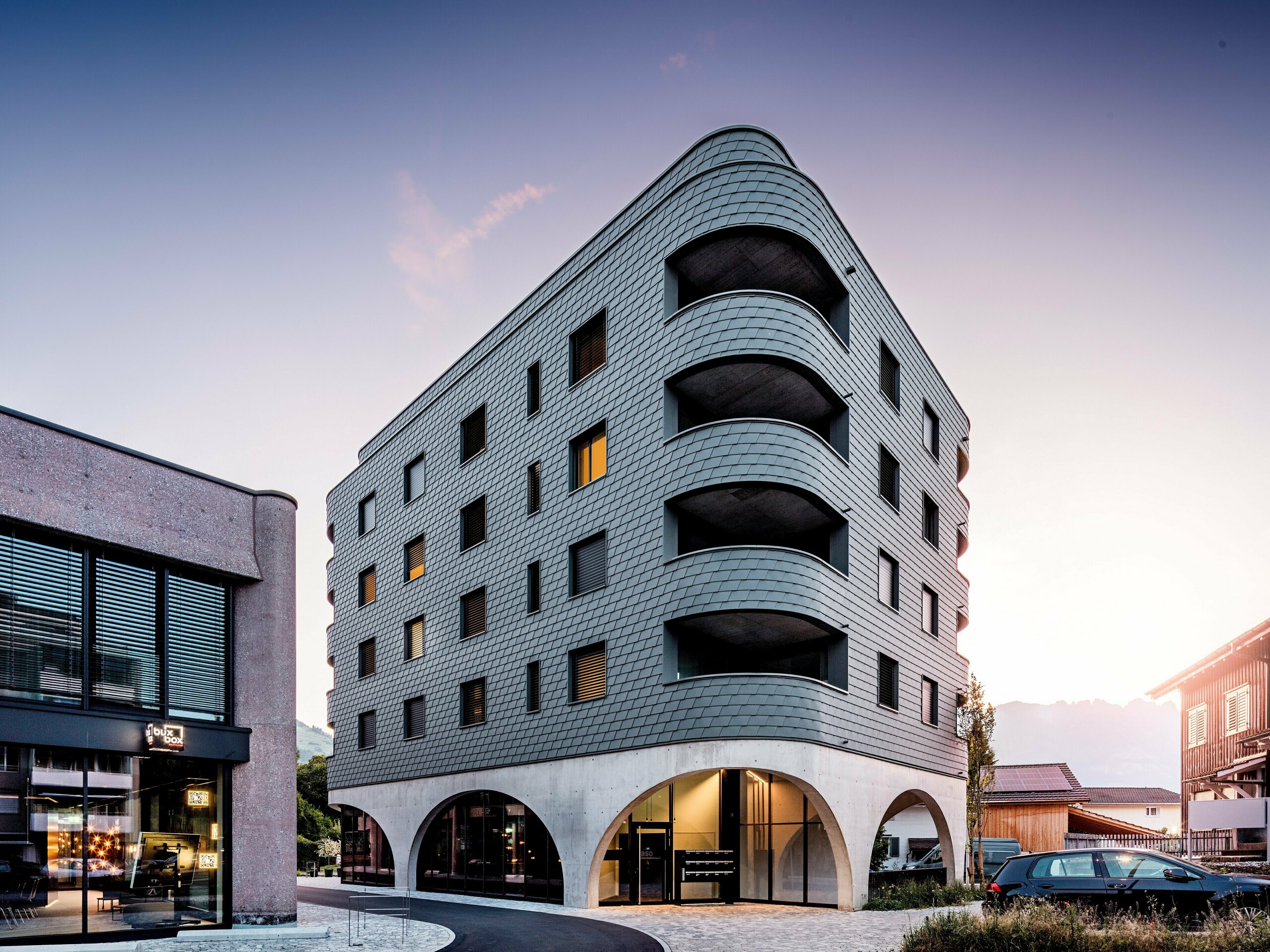 Un immeuble moderne à usage mixte à Buchs, à la forme triangulaire arrondie saisissante et à la façade continue en bardage PREFA gris souris P.10. L'enveloppe en aluminium, aux allures d'écailles, épouse la structure du bâtiment comme un reptile, soulignant ses courbes fluides et ses balcons profondément encastrés. Au-dessus d'un socle massif aux élégantes arcades en béton, la façade PREFA, légère et résistante aux intempéries, se distingue par son expressivité. La pose précise des bardeaux d'aluminium garantit une enveloppe homogène, durable et nécessitant peu d'entretien, tout en assurant une forte identité visuelle.