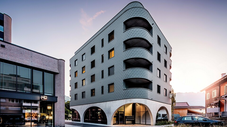 Un immeuble moderne à usage mixte à Buchs, à la forme triangulaire arrondie saisissante et à la façade continue en bardage PREFA gris souris P.10. L'enveloppe en aluminium, aux allures d'écailles, épouse la structure du bâtiment comme un reptile, soulignant ses courbes fluides et ses balcons profondément encastrés. Au-dessus d'un socle massif aux élégantes arcades en béton, la façade PREFA, légère et résistante aux intempéries, se distingue par son expressivité. La pose précise des bardeaux d'aluminium garantit une enveloppe homogène, durable et nécessitant peu d'entretien, tout en assurant une forte identité visuelle.