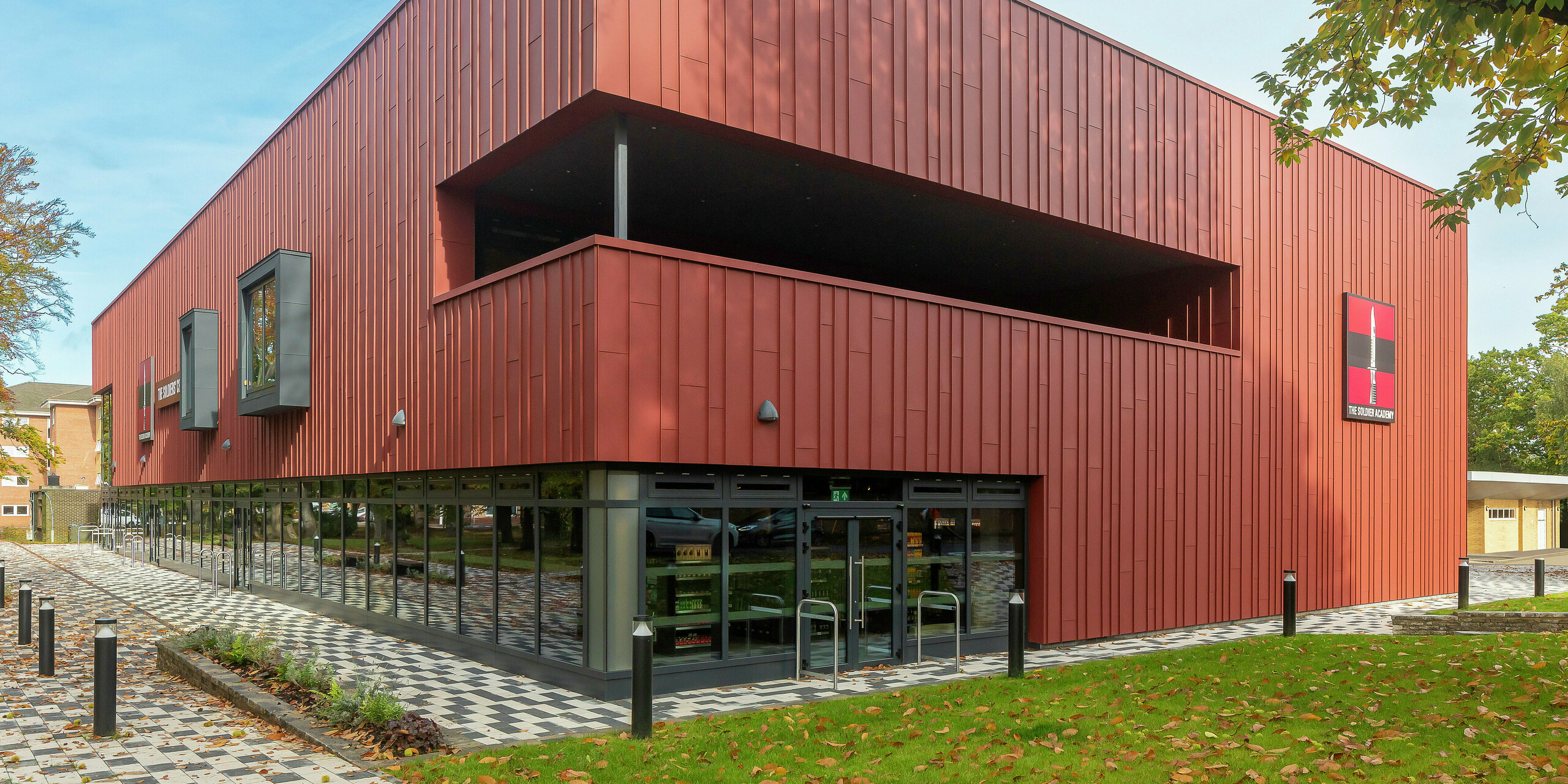 Architecture contemporaine avec PREFALZ : Le Soldiers' Centre de Pirbright impressionne par sa façade moderne à joint debout en PREFALZ en P.10 rouge oxyde. Les vastes baies vitrées du rez-de-chaussée créent transparence et ouverture, tandis que les cadres de fenêtres en P.10 anthracite ajoutent des accents élégants. Un concept de construction durable avec des produits en aluminium PREFA durables pour les normes de conception et de qualité les plus élevées.