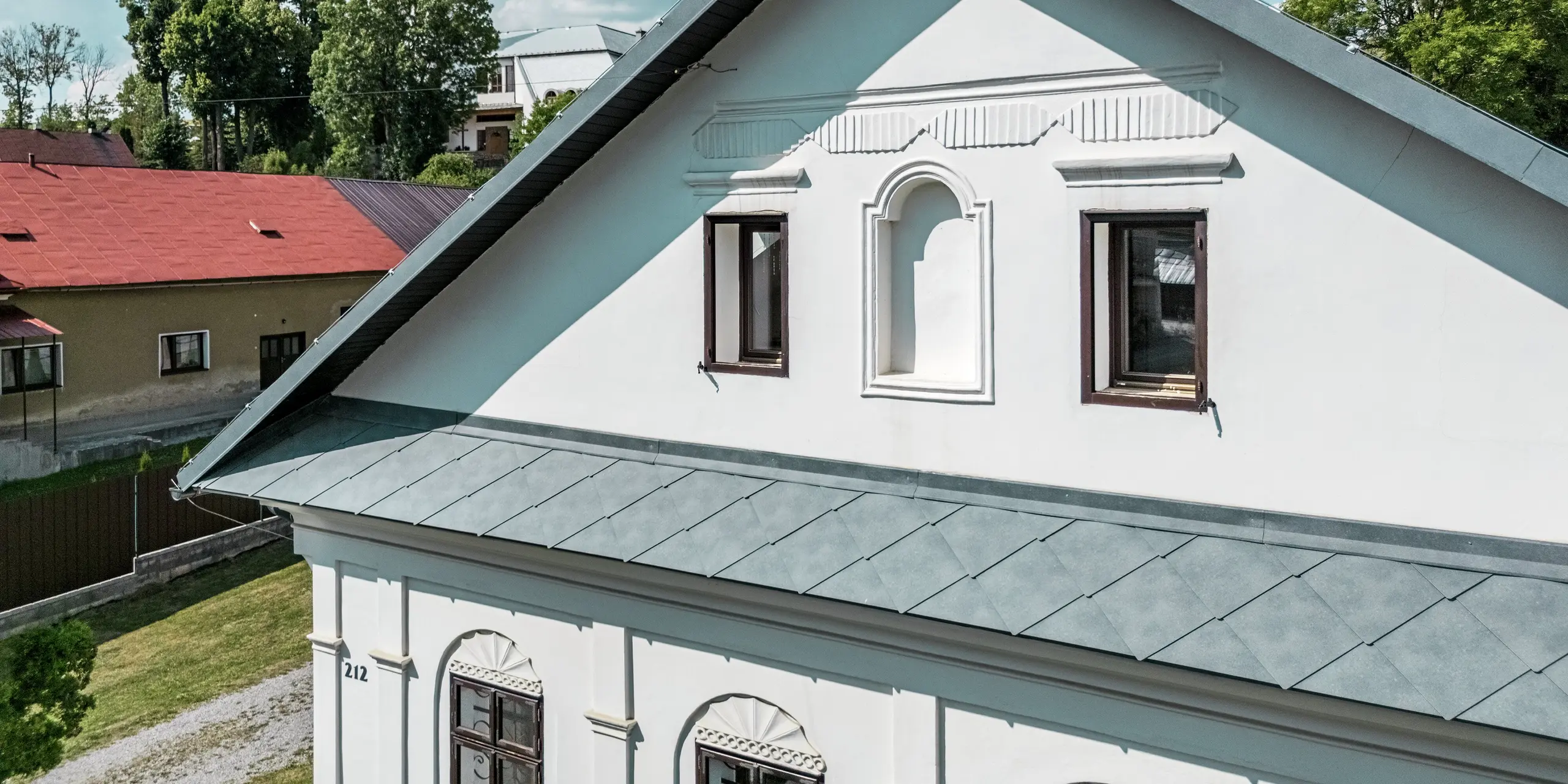 Le gros plan révèle la toiture d'une maison d'artisan à Klin, en Slovaquie, recouverte de losanges PREFA 44 x 44 cm, coloris Gris Pierre P.10. L'harmonieuse alliance entre la toiture moderne et la façade historique souligne le caractère du bâtiment. L'angle prononcé du toit est particulièrement saisissant et s'intègre parfaitement à l'architecture traditionnelle. Les tuiles en aluminium, assorties à la couleur de la toiture, offrent non seulement un aspect esthétique, mais sont également résistantes à la corrosion, légères et durables. Cette solution apporte une amélioration durable, fonctionnelle et esthétique à la rénovation de la maison.