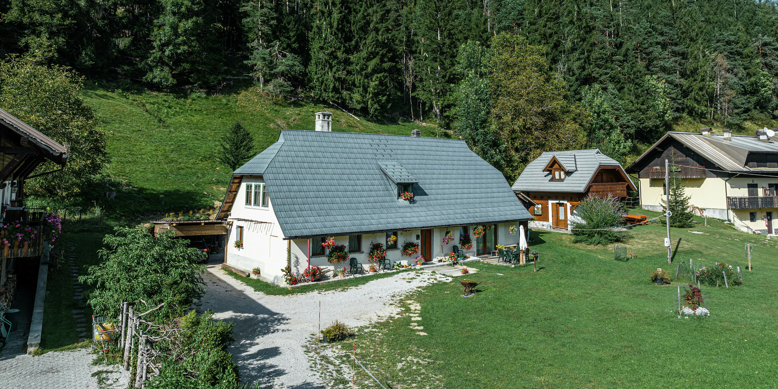 Vue aérienne d'une maison familiale idyllique à Tarvisio, en Italie, protégée par des tuiles PREFA durables depuis 1972. Le grand toit en aluminium résistant aux intempéries s'intègre harmonieusement dans le paysage alpin verdoyant. La façade joliment décorée de fleurs et la terrasse chaleureuse confèrent à la maison une atmosphère accueillante. Entourée de maisons traditionnelles aux façades en bois et d'une forêt dense de conifères, la propriété offre un mélange parfait de proximité avec la nature et de stabilité architecturale.