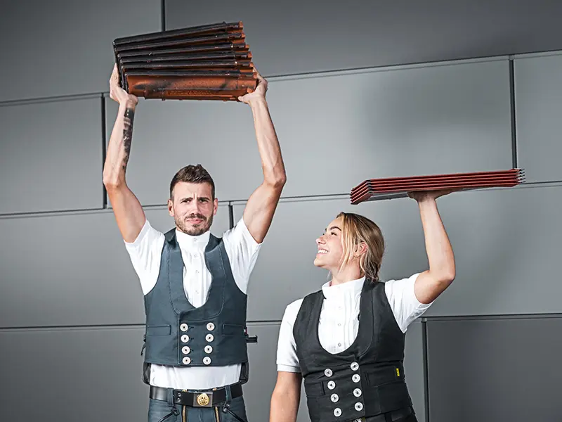 Sur la photo, on voit deux personnes tenant chacune une pile de matériaux de couverture. La personne à gauche tient une pile de tuiles au-dessus de sa tête, tandis que celle à droite tient une pile de panneaux de toiture en aluminium, également au-dessus de sa tête. Toutes deux portent une tenue traditionnelle d'artisan, avec un gilet et une chemise blanche. L'arrière-plan est moderne et minimaliste, avec des murs gris. Les deux personnes semblent concentrées et sourient.
