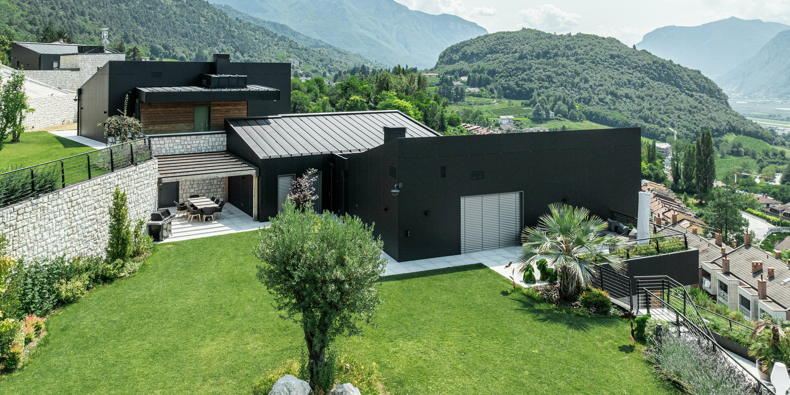 Complexe résidentiel moderne à flanc de colline avec une vue imprenable sur le paysage montagneux de Trente. La façade en panneaux composites en aluminium PREFA en P.10 noir assure une enveloppe de bâtiment élégante et résistante aux intempéries. Le toit PREFALZ en P.10 Noir complète le design minimaliste et offre une durabilité maximale. Un paysage de jardin bien entretenu avec des murs en pierre naturelle s'intègre harmonieusement dans l'environnement et souligne le caractère résidentiel exclusif.