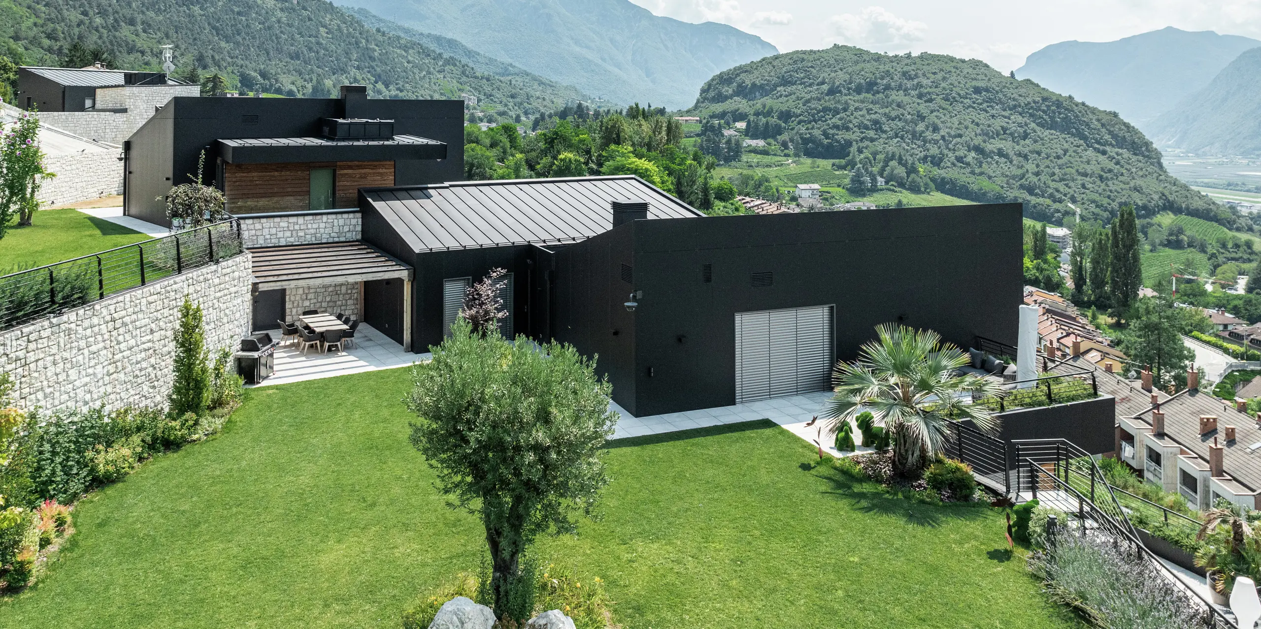 Complexe résidentiel moderne à flanc de colline avec une vue imprenable sur le paysage montagneux de Trente. La façade en panneaux composites en aluminium PREFA en P.10 noir assure une enveloppe de bâtiment élégante et résistante aux intempéries. Le toit PREFALZ en P.10 Noir complète le design minimaliste et offre une durabilité maximale. Un paysage de jardin bien entretenu avec des murs en pierre naturelle s'intègre harmonieusement dans l'environnement et souligne le caractère résidentiel exclusif.