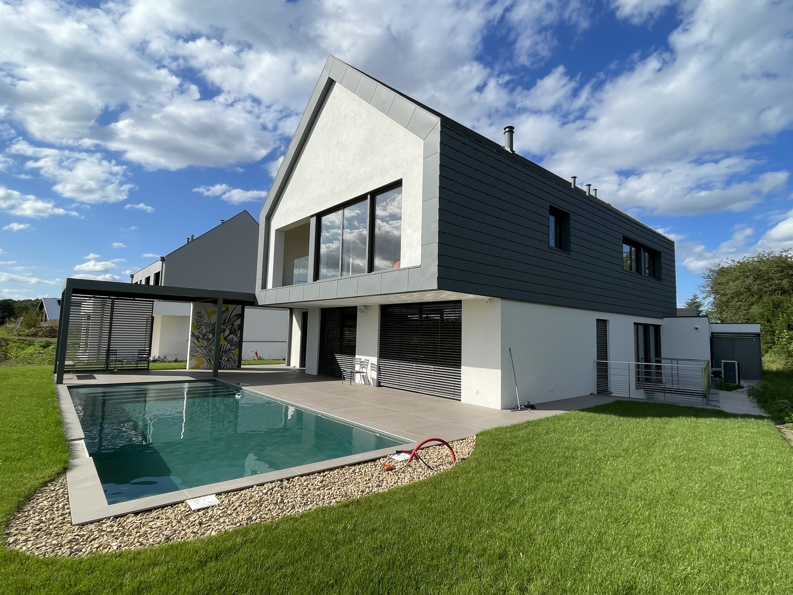 Vue d'ensemble de cette nouvelle maison individuelle dont la toiture et la façade contemporaine ont été recouvertes de bardeaux en aluminium PREFA. On aperçoit une jolie piscine en premier plan.