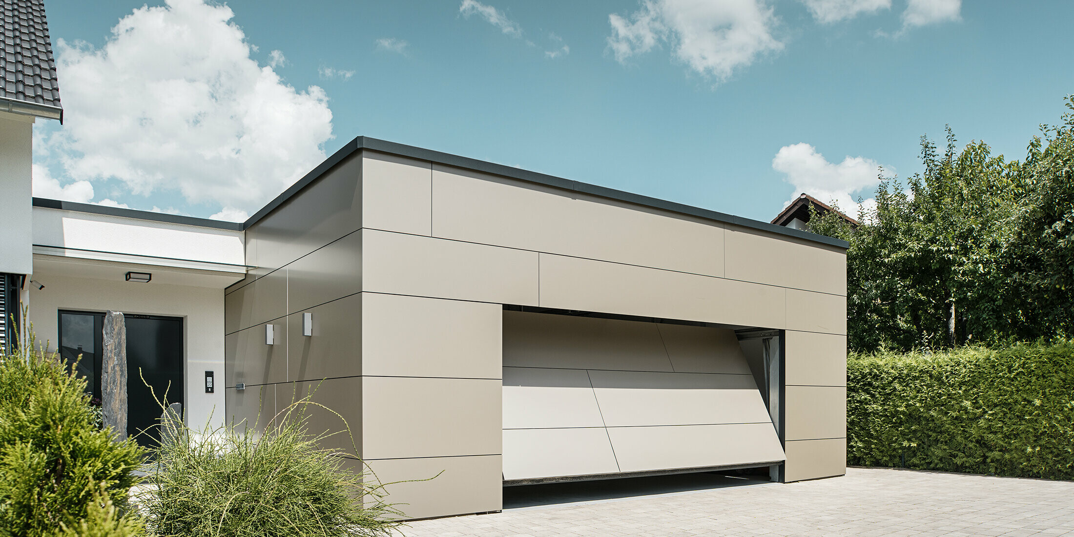 Gesamtansicht einer mit PREFABOND Aluminium Verbundplatten in Bronze verkleideten Garage. Das automatische Garagentor öffnet sich gerade und ist bereits ein Stück geöffnet. Direkt neben der Garage befindet sich der Hauseingang mit anthrazitfarbener Tür und weißer Putzfassade.