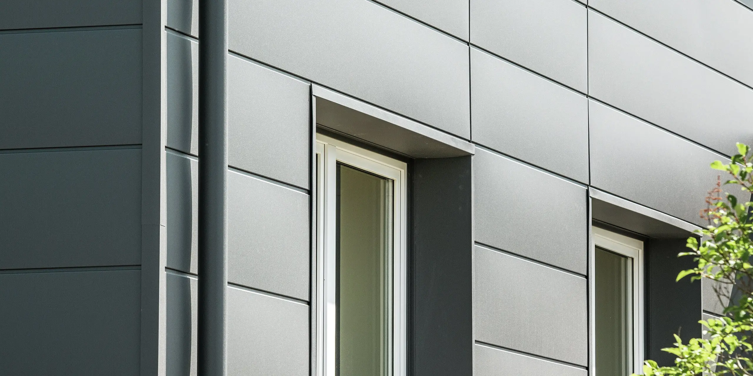 Vue détaillée d'une façade en Sidings PREFA couleur P.10 gris sombre. L'intégration harmonieuse des cadres de fenêtres et la pose précise des panneaux confèrent à la façade une esthétique à la fois harmonieuse et fonctionnelle. La surface résistante protège durablement le bâtiment des intempéries et garantit un entretien minimal. L'image illustre la parfaite alliance du design et de la durabilité, la façade séduisant par son allure épurée et contemporaine.
