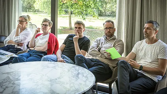 Réunion du jury de PREFARENZEN 2027 dans une salle de séminaire lumineuse : plusieurs architectes et experts en design sont assis attentivement devant des panneaux d’affichage bleus présentant des projets architecturaux intégrant l’aluminium PREFA. De grandes fenêtres laissent entrer la lumière naturelle et offrent une vue sur un jardin verdoyant. Des livres d’architecture, des carnets et des stylos sont disposés sur les tables rondes. La scène témoigne d’une discussion intense, d’une évaluation créative du design et de la fonctionnalité, et de l’inspiration que les produits en aluminium PREFA offrent à travers des toitures et des façades durables et esthétiques.
