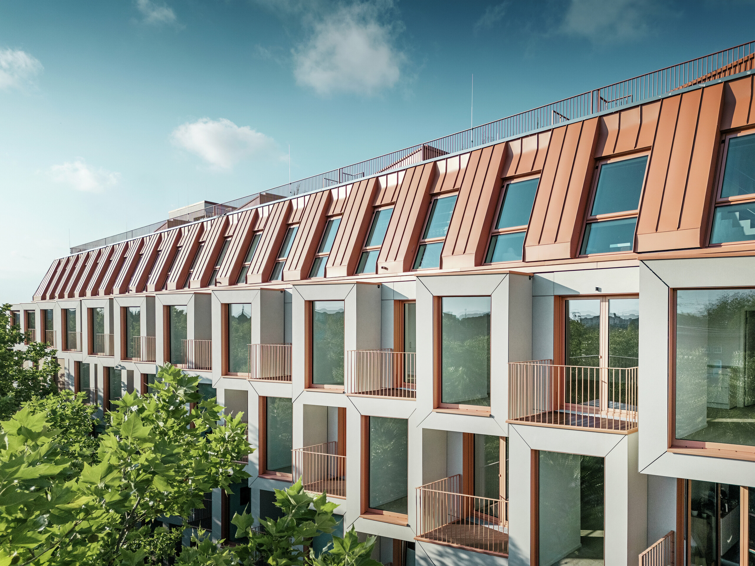 Vue de la façade du projet résidentiel Van B à Munich avec le toit en aluminium PREFALZ remarquable dans la couleur spéciale IGP Orange HDP de PREFA. La couverture de toit moderne allie élégance esthétique et précision technique et offre une protection durable contre les éléments. Les baies vitrées de conception cubique avec de généreuses surfaces vitrées et des balcons délicats créent une interaction harmonieuse entre lumière, espace et fonctionnalité - idéale pour des concepts de vie sophistiqués dans un environnement urbain.