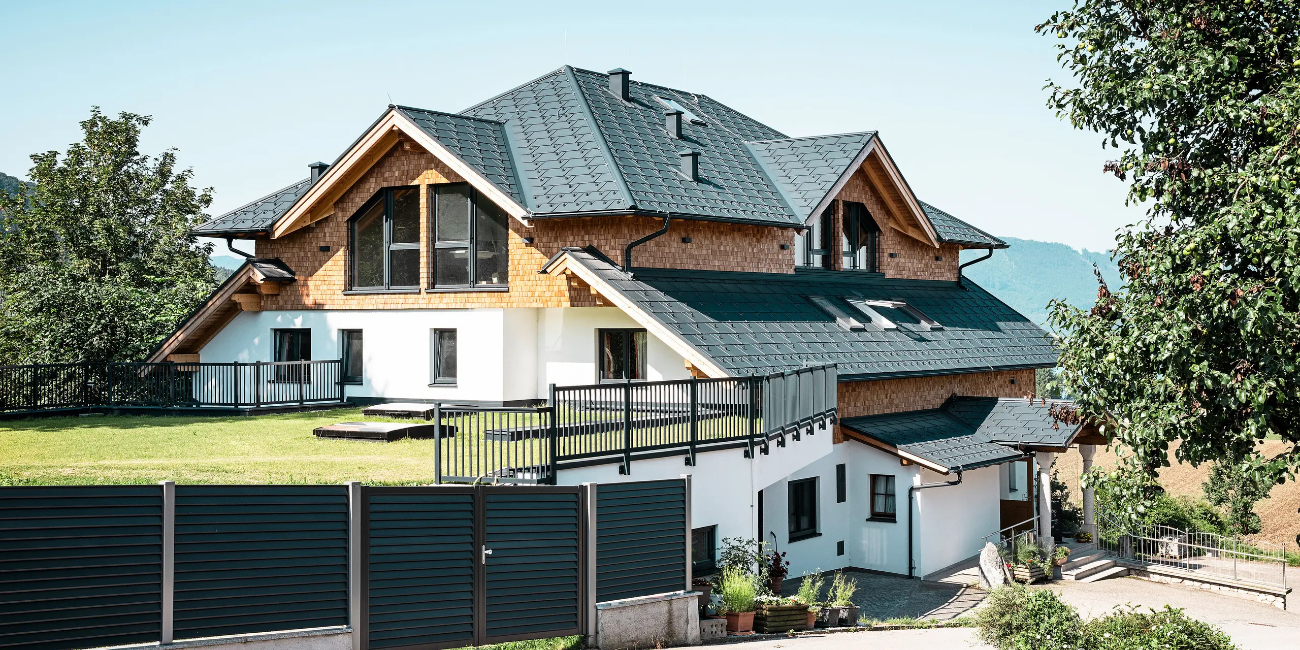 Vue latérale du café-restaurant Zenz'n Stub'n à Schörfling am Attersee avec un toit moderne en aluminium composé de panneaux de toiture PREFA R.16 avec revêtement de haute qualité P.10 anthracite. Le toit durable PREFA allie esthétique et fonctionnalité et offre une protection optimale. L'accès au jardin et l'intégration harmonieuse du bâtiment dans l'environnement naturel soulignent le design intemporel.