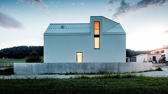 L'image présente une superbe maison individuelle à Freistadt, qui impressionne par ses lignes épurées et son architecture minimaliste. La façade d'un blanc immaculé, réalisée en aluminium plié PREFALZ, forme une structure géométrique distinctive qui confère au bâtiment une allure à la fois moderne et intemporelle. Le toit à forte pente, intégrant une lucarne, et les grandes fenêtres assurent une luminosité naturelle abondante, rendant l'intérieur spacieux et accueillant. L'utilisation de l'aluminium PREFALZ garantit non seulement une esthétique durable, mais aussi une excellente résistance aux intempéries et une grande longévité, faisant de cette maison un havre de fonctionnalité et de durabilité. La clarté du design et l'élégance des détails contribuent à une solution à la fois raffinée et pratique pour l'architecture résidentielle contemporaine.