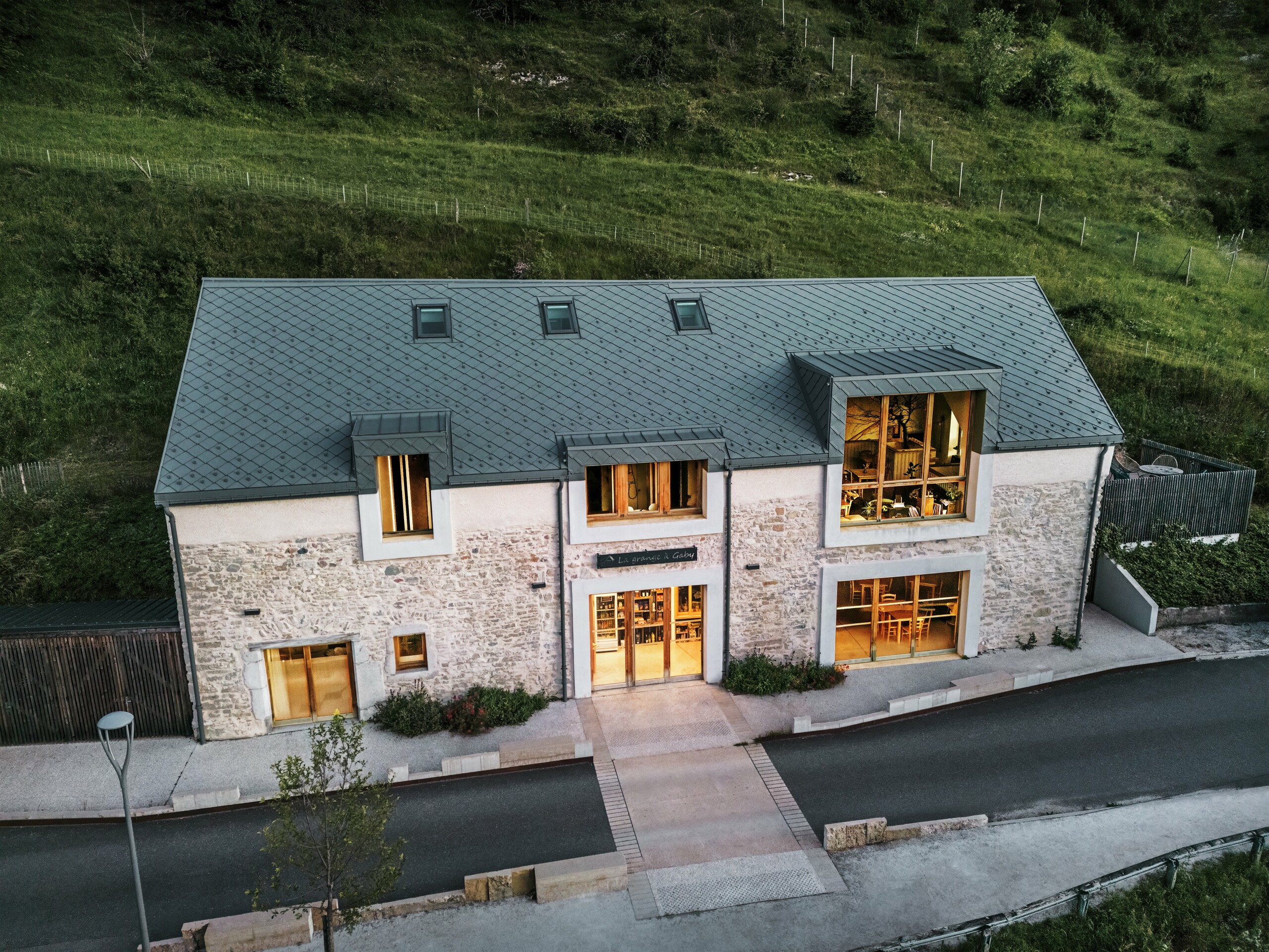 Vue nocturne de la ferme restaurée "La Grange à Gaby" à Engins, France. Le bâtiment combine la maçonnerie en pierre traditionnelle avec un toit moderne en aluminium PREFA composé de losanges de toiture de 29 × 29 cm. Les grandes fenêtres et les intérieurs illuminés soulignent l’atmosphère accueillante du nouveau centre culturel.