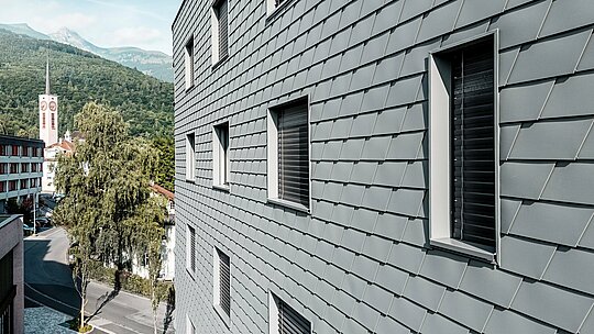 Vue détaillée de la façade de la Bernerhaus à Buchs, revêtue de bardages PREFA P.10 Gris souris, posés avec précision. La surface en aluminium, à l'aspect d'écailles, crée un motif fin et dynamique et souligne les ouvertures de fenêtres aux embrasures profondes. Léger et résistant aux intempéries, l'aluminium PREFA garantit une enveloppe de bâtiment homogène, indéformable et nécessitant peu d'entretien. Sur fond de paysage alpin, cette façade métallique haut de gamme devient un élément architectural moderne et intemporel.