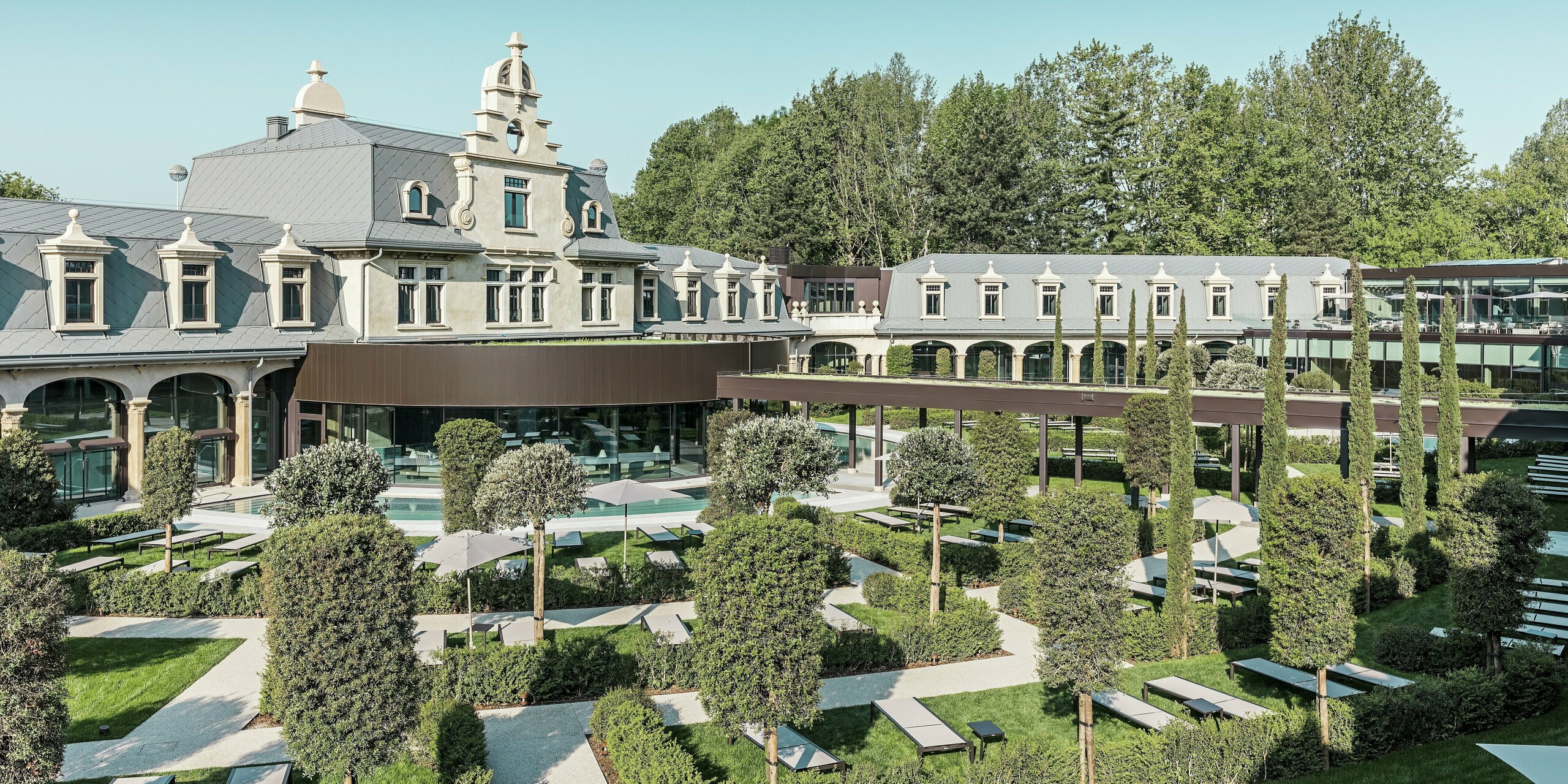 Le parc thermal « De Montel – Terme Milano », soigneusement restauré, présente un jardin paysager moderne avec des allées linéaires, des espaces détente et des plantes méditerranéennes. L'élégante toiture en losanges PREFA 44 × 44 P.10 gris souris souligne l'architecture historique, tandis que des éléments vitrés modernes et une verrière végétalisée créent un lien avec l'utilisation contemporaine.