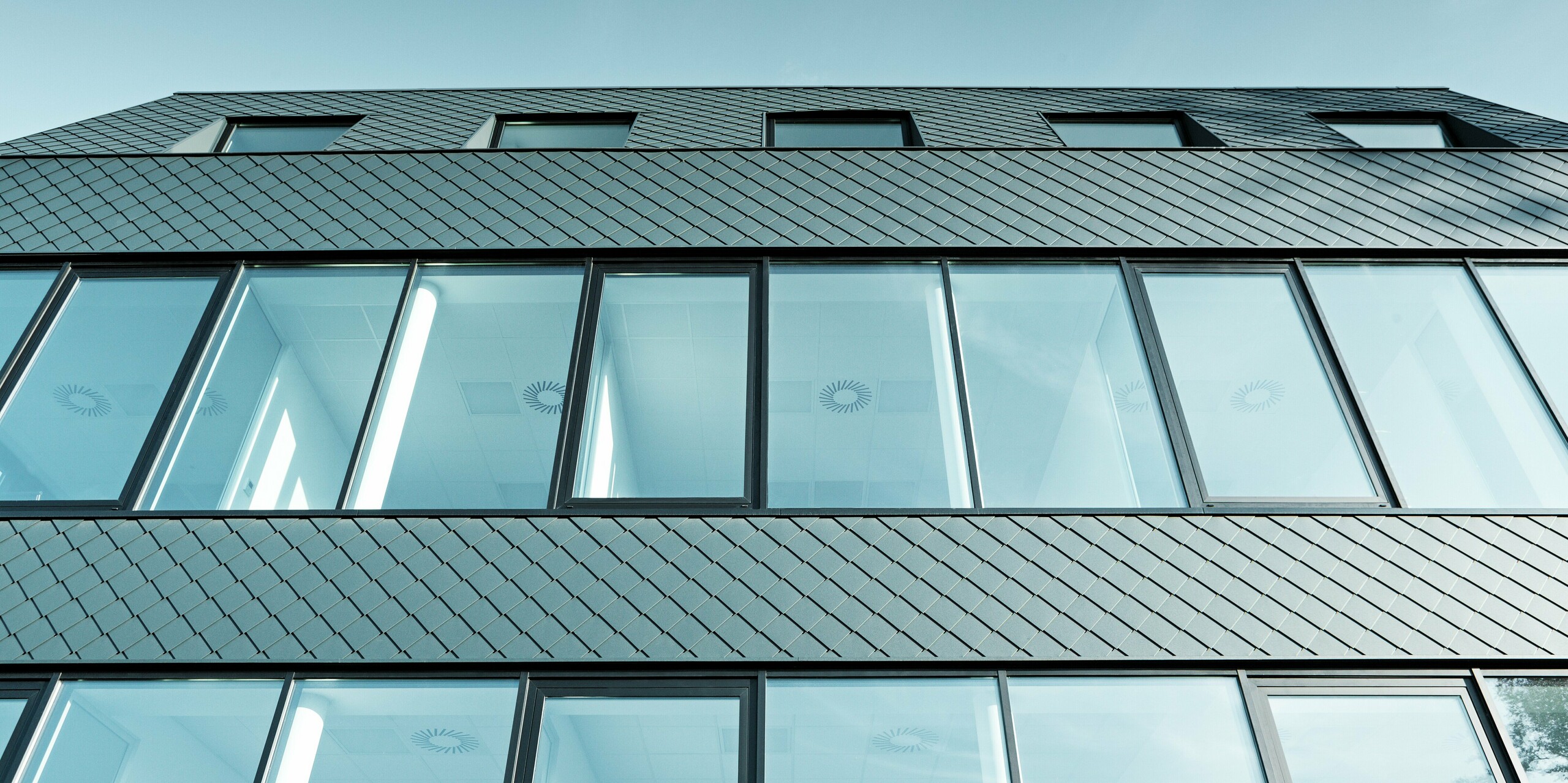 Dynamische Aufwärtsansicht des modernen Fassadenzubaus am Kasteeltje Heizel: Die anthrazitfarbenen PREFA Wandrauten bilden in präziser Rautenstruktur einen markanten Kontrast zu den großflächigen Glasfronten. Das Spiel von Licht und Spiegelungen verstärkt die moderne Eleganz und unterstreicht die Kombination aus langlebiger Aluminiumfassade und klarer Architektur.