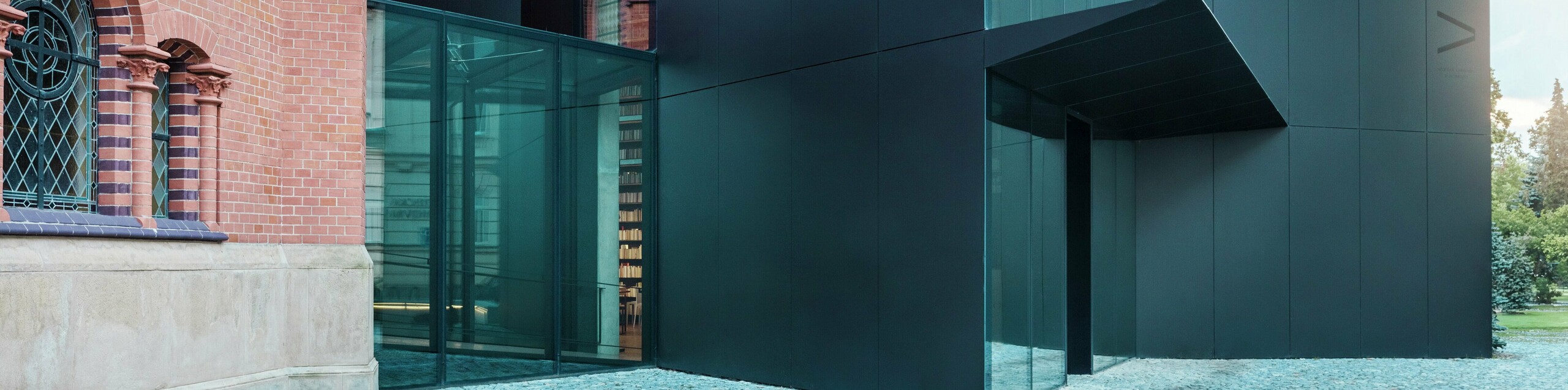L'image présente l'extension moderne d'une bibliothèque, élégamment reliée à l'église rouge historique d'Olomouc. Ce nouveau bâtiment remarquable impressionne par sa façade en panneaux composites en aluminium PREFABOND noirs, aux formes géométriques épurées et aux détails précis. Les parois vitrées offrent une vue transparente sur l'intérieur, qui se fond harmonieusement dans le paysage historique. Le jeu des matériaux modernes et des éléments architecturaux classiques crée un lien visuel captivant entre ancien et nouveau. L'utilisation innovante des panneaux PREFABOND garantit non seulement une esthétique contemporaine, mais aussi une grande durabilité et une résistance à toute épreuve, préparant ainsi le bâtiment aux enjeux futurs.