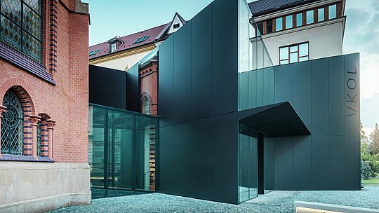 L'image présente l'extension moderne d'une bibliothèque, élégamment reliée à l'église rouge historique d'Olomouc. Ce nouveau bâtiment remarquable impressionne par sa façade en panneaux composites en aluminium PREFABOND noirs, aux formes géométriques épurées et aux détails précis. Les parois vitrées offrent une vue transparente sur l'intérieur, qui se fond harmonieusement dans le paysage historique. Le jeu des matériaux modernes et des éléments architecturaux classiques crée un lien visuel captivant entre ancien et nouveau. L'utilisation innovante des panneaux PREFABOND garantit non seulement une esthétique contemporaine, mais aussi une grande durabilité et une résistance à toute épreuve, préparant ainsi le bâtiment aux enjeux futurs.