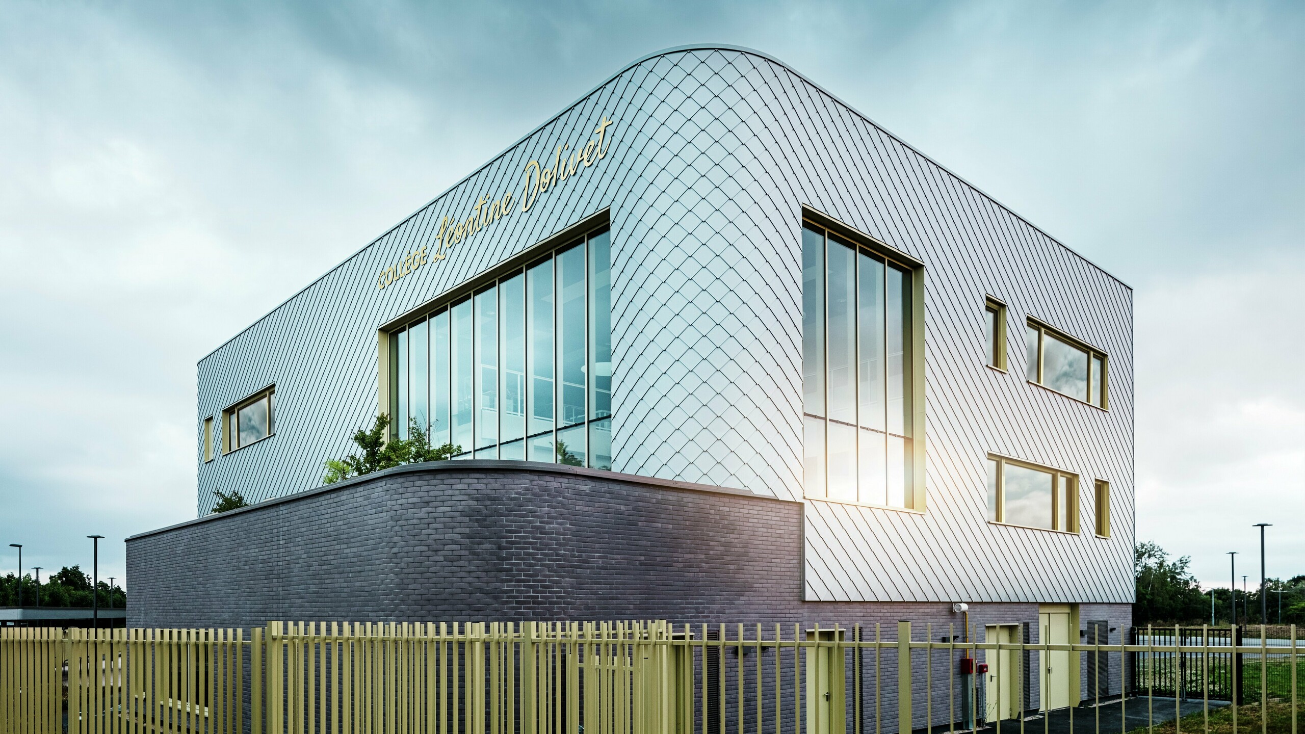 La photo montre l'angle remarquable du Collège Léontine Dolivet, avec sa façade incurvée en aluminium composée de losanges de façade PREFA argent métallisé. De larges surfaces vitrées reflètent la lumière, tandis que des encadrements de fenêtres jaunes apportent une touche d'originalité. Au premier plan, une clôture claire longe le bâtiment, reposant sur un socle de briques sombres. Le ciel est couvert.