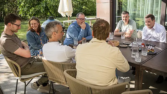 PREFARENZEN 2027 : Dialogue d'experts dans la terrasse ensoleillée d'une maison de vigneron : architectes et designers, absorbés par leurs discussions, sont assis autour d'une grande table, tandis que verres et amuse-bouches sont disposés. La scène, baignée de verdure et à l'ombre des arbres, illustre une interaction animée et une attention soutenue. En arrière-plan, les produits en aluminium PREFA représentent des toitures et des façades à la fois durables et esthétiques. L'image montre comment PREFA conjugue design, fonctionnalité et durabilité, stimulant ainsi les échanges créatifs entre architectes.