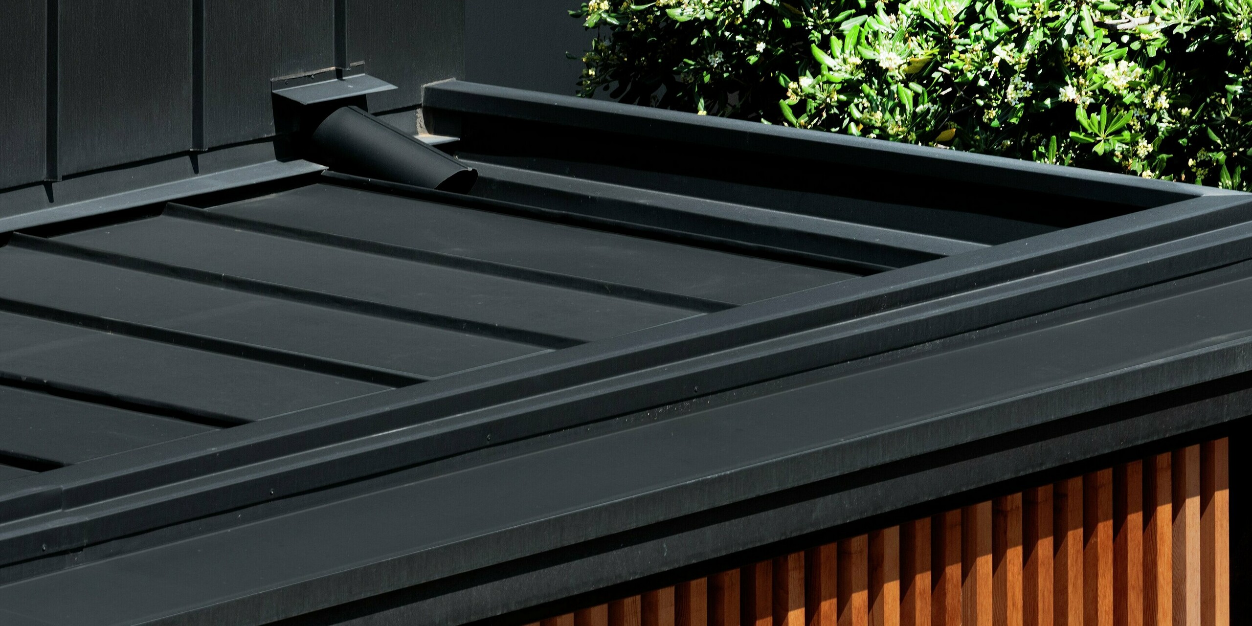 Vue détaillée d'une maison moderne à Cattolica : élégante façade en lattes de bois associée à une toiture et une façade PREFALZ gris noir. La surface en aluminium PREFA offre durabilité, protection contre la corrosion et lignes épurées – idéale pour une architecture contemporaine alliant charme naturel et haute résistance. Le système d'évacuation des eaux pluviales dissimulé est visible sur l'image.