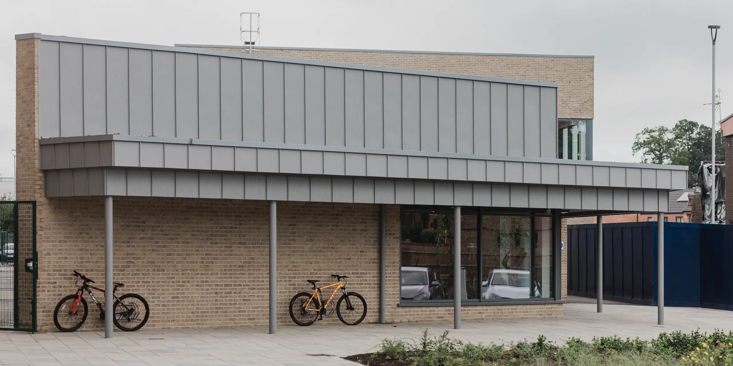 Le Northern Regional College de Ballymena dispose d'un parking à vélos moderne, doté d'un vaste auvent en aluminium PREFALZ gris quartz. Les joints debout, à structure verticale, se prolongent avec précision au-dessus du toit, créant un contraste visuel saisissant avec la façade en briques. Colonnes élancées, murs de briques claires, vélos stationnés et cour ouverte soulignent le caractère convivial du campus. L'aluminium PREFA séduit par sa durabilité, sa résistance aux intempéries et son aspect sobre et élégant au quotidien.