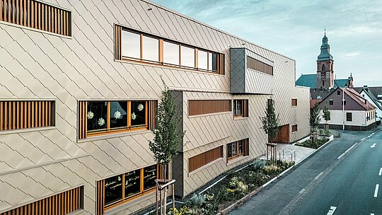 La vue latérale de la crèche d'Alzenau révèle un bâtiment hybride et moderne qui contraste fortement avec le paysage historique de la ville, dominé par l'église gothique tardive du château. La façade, composée de losanges de façade 44x44 PREFA couleur bronze, dessine un motif élégant et rythmé et se distingue avec assurance dans son environnement, sans pour autant l'imposer. De larges ouvertures vitrées, dotées de persiennes verticales en bois, et la végétation structurée qui longe le bâtiment soulignent le concept de conception durable. L'alliance d'une façade en aluminium innovante, d'une construction écologique et d'une approche urbanistique respectueuse de l'environnement fait de ce projet un exemple remarquable d'architecture responsable, qui fait le lien entre tradition et modernité.