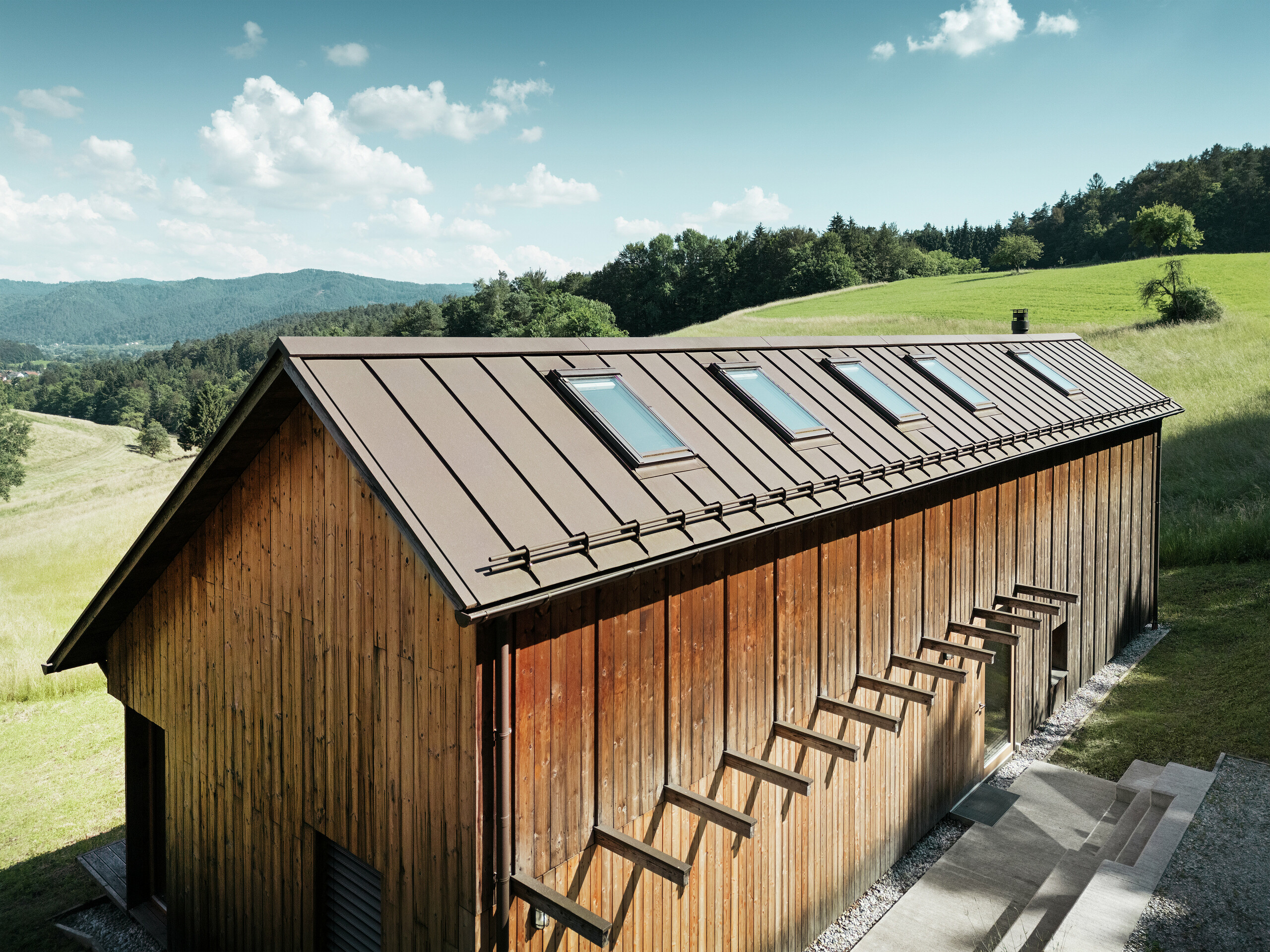 Maison en bois à l’architecture clairement structurée, avec un toit en aluminium PREFALZ de haute qualité en P.10 brun. Ce toit PREFA, nécessitant peu d’entretien, séduit par la précision de ses lignes, sa grande résistance aux UV et à la corrosion, ainsi que par l’intégration parfaite des fenêtres de toit. L’association d’une façade en bois durable et d’une couverture à joint debout longue durée s’intègre harmonieusement dans le paysage vallonné idyllique. Le toit à deux pans asymétrique est équipé de cinq fenêtres de toit posées avec précision, apportant de la lumière à l’intérieur.