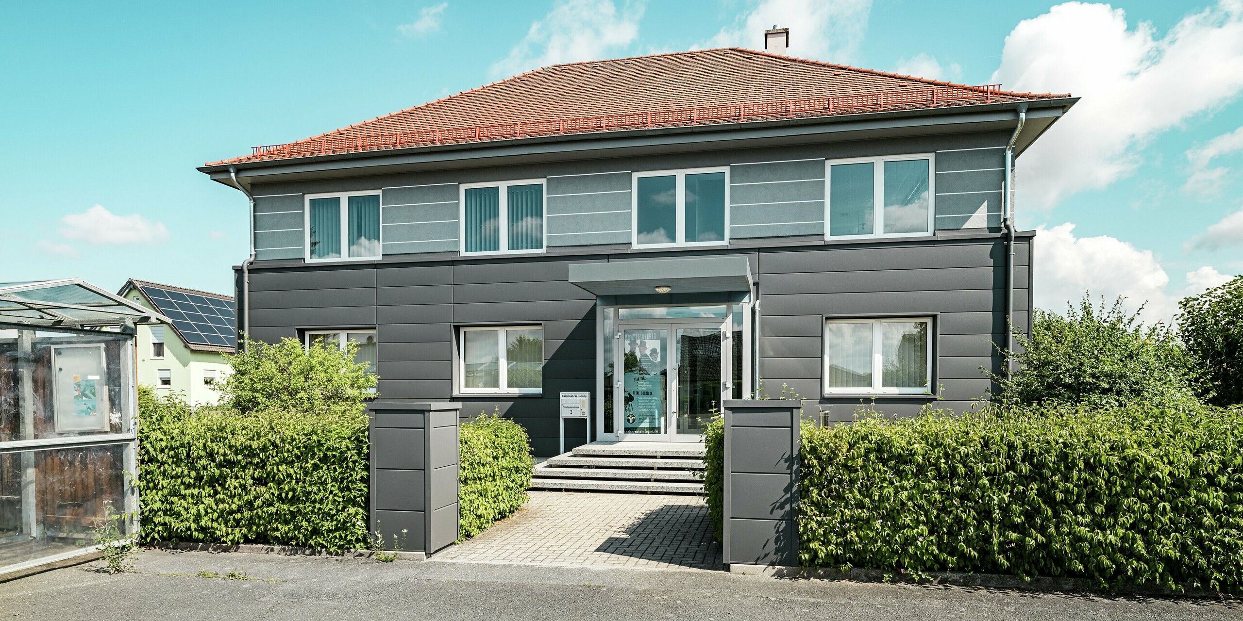 La façade rénovée de la guilde des ramoneurs de Bindlach est revêtue de panneaux PREFA de haute qualité, couleur Gris sombre P.10. Ces panneaux d'aluminium à la structure apparente confèrent au bâtiment une allure contemporaine et élégante. Le design moderne met en valeur l'entrée, tandis que les panneaux s'intègrent harmonieusement à la façade. Ce matériau est non seulement durable et facile d'entretien, mais il offre également une protection pérenne et se fond parfaitement dans son environnement. La verdure et le ciel dégagé créent un magnifique contraste avec le bâtiment gris et ses tuiles rouges sur le toit à quatre pans.