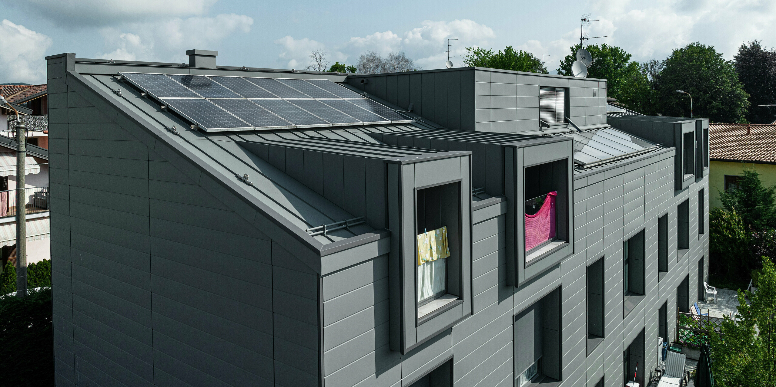 Vue aérienne du toit d'un complexe résidentiel moderne à Faloppio, en Italie, avec un revêtement robuste en aluminium à joint debout et un système photovoltaïque durable. La façade en bardage PREFA et le toit métallique offrent non seulement des points forts visuels, mais également une excellente résistance aux intempéries et une source d'énergie durable grâce aux modules photovoltaïques. La combinaison de produits PREFA fonctionnels et esthétiques garantit une solution durable et économe en énergie.