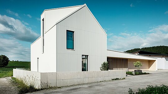 Cette maison individuelle moderne à Freistadt est présentée sous un angle dynamique, mettant en valeur ses lignes épurées et son architecture remarquable. Le bardage en aluminium blanc pur PREFALZ s'étend harmonieusement sur toute la façade et la toiture, conférant à la maison une esthétique à la fois moderne et intemporelle. Parmi ses éléments architecturaux caractéristiques, on retrouve le toit à forte pente, les grandes fenêtres et la lucarne distinctive, tandis que les rainures verticales de la façade apportent texture et profondeur au bâtiment. Le contraste avec l'environnement naturel, ainsi que les élégants éléments en bois de l'entrée et de la porte de garage, parachèvent l'ensemble. PREFALZ offre une surface particulièrement durable, facile d'entretien et résistante aux intempéries. Ainsi, la maison est non seulement esthétique, mais aussi fonctionnelle et durable.