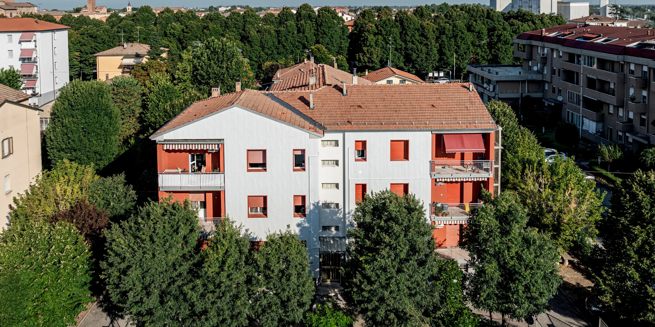 Vue panoramique sur le complexe résidentiel Alba B, niché dans un écrin de verdure urbain : le bâtiment rénové se distingue nettement des toits de tuiles environnants grâce à sa façade lumineuse PREFA, composée de panneaux d'aluminium Siding.X. La pose verticale des panneaux crée une surface finement texturée, tandis que les encadrements de fenêtres et les balcons rouges apportent une touche d'originalité. Entouré d'une végétation dense, l'ensemble paraît dynamique et moderne. La façade en aluminium PREFA séduit par sa durabilité, sa résistance aux intempéries et son faible besoin d'entretien.