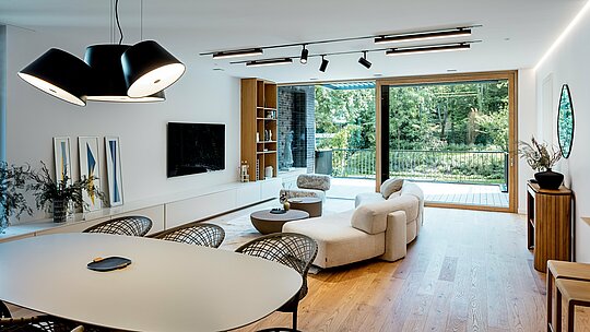 La photographie révèle le séjour et la salle à manger baignés de lumière d'une villa moderne et intergénérationnelle à Budapest. De larges baies vitrées ouvrent la pièce sur une terrasse verdoyante, tandis que le parquet clair, le mobilier design élégant et un éclairage harmonieux créent une atmosphère chaleureuse. L'agencement ouvert relie avec fluidité la cuisine, la salle à manger et le séjour. La qualité de l'aménagement intérieur s'accorde parfaitement avec les lignes épurées du bâtiment, dont la structure extérieure est caractérisée par des éléments en aluminium PREFALZ P.10 anthracite, gage de durabilité. L'espace dégage une impression d'espace, de modernité et de confort optimal.