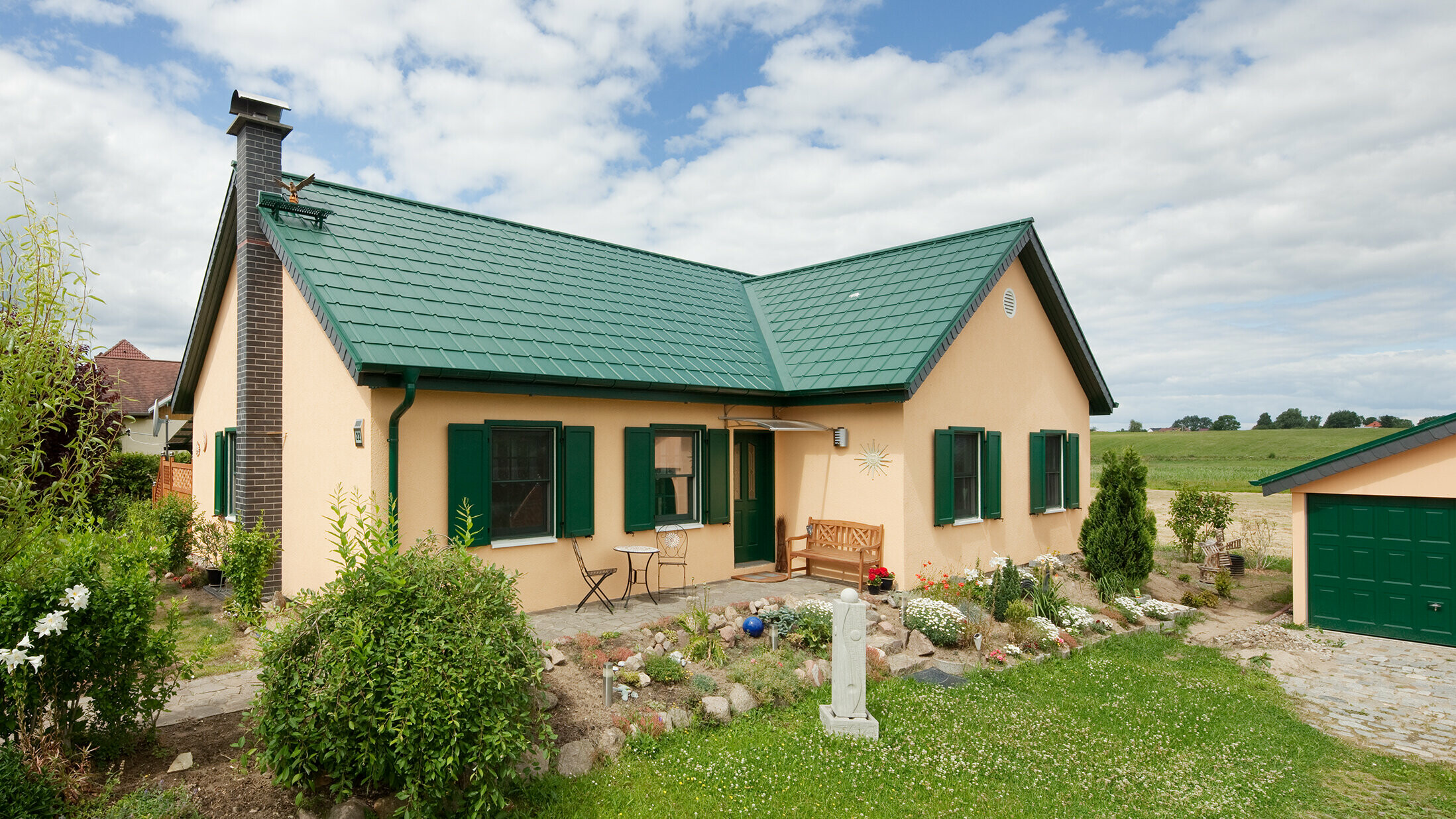 Toit en bâtière élargi ; toit en bâtière en forme de L ; avec la tuile PREFA en P.10 vert mousse ; jolie petite maison de campagne avec des volets verts assortis à la toiture PREFA.