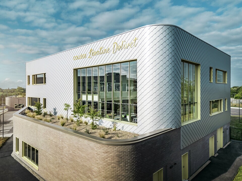 La photo révèle un angle remarquable du collège Léontine Dolivet à Cesson-Sévigné. La façade est composée de losanges PREFA argent métallisé, agencés en diagonale et jouant avec la lumière. La forme incurvée de l'étage supérieur est particulièrement frappante et confère au bâtiment une allure douce et moderne. De larges baies vitrées offrent des vues sur l'intérieur, tandis que les encadrements de fenêtres jaunes apportent des touches de dynamisme. Au premier plan, un espace planté de gravier et d'arbustes s'intègre harmonieusement à l'architecture. Le ciel légèrement couvert accentue encore la brillance des surfaces métalliques.