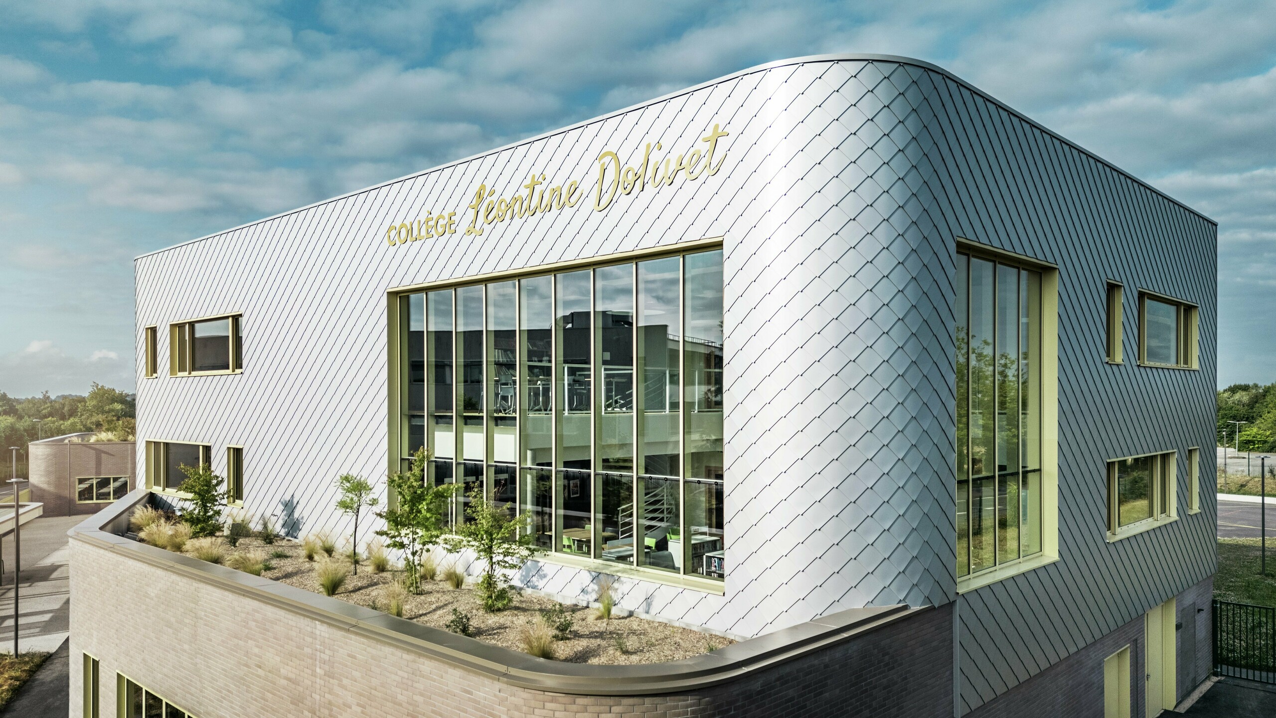 La photo révèle un angle remarquable du collège Léontine Dolivet à Cesson-Sévigné. La façade est composée de losanges PREFA argent métallisé, agencés en diagonale et jouant avec la lumière. La forme incurvée de l'étage supérieur est particulièrement frappante et confère au bâtiment une allure douce et moderne. De larges baies vitrées offrent des vues sur l'intérieur, tandis que les encadrements de fenêtres jaunes apportent des touches de dynamisme. Au premier plan, un espace planté de gravier et d'arbustes s'intègre harmonieusement à l'architecture. Le ciel légèrement couvert accentue encore la brillance des surfaces métalliques.