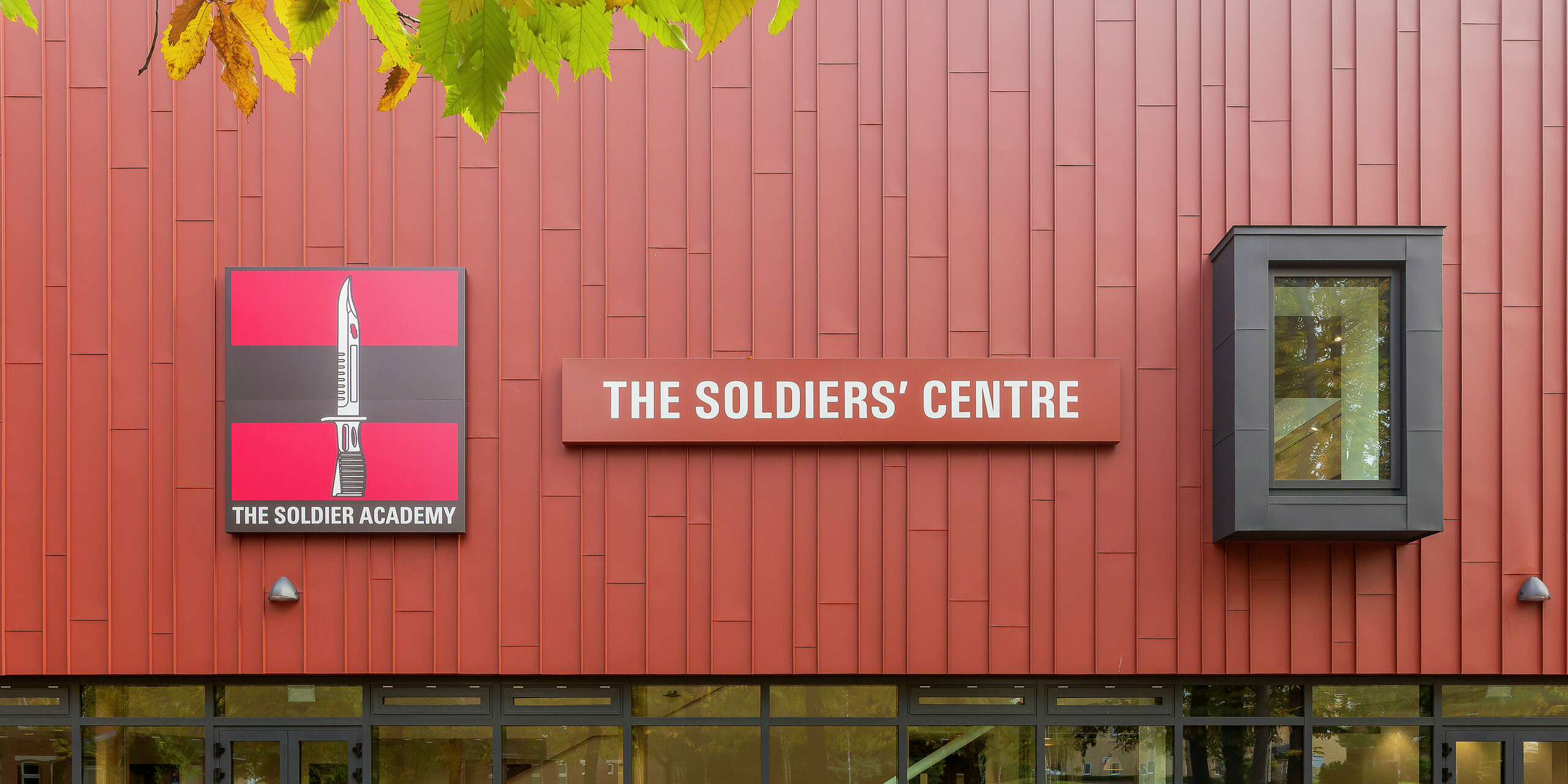 Bâtiment social construit spécialement pour l'armée avec façade PREFALZ : La toiture à joint debout traitée avec précision en P.10 rouge oxyde confère au bâtiment une apparence remarquable. La zone d'entrée impressionne par ses généreuses façades vitrées qui offrent luminosité et transparence. Le logo « Soldier Academy » et le panneau de bâtiment accrocheur créent des accents visuels clairs. Les cadres de fenêtre en P.10 anthracite complètent harmonieusement le design. Produits en aluminium PREFA de haute qualité pour une architecture durable et esthétique.