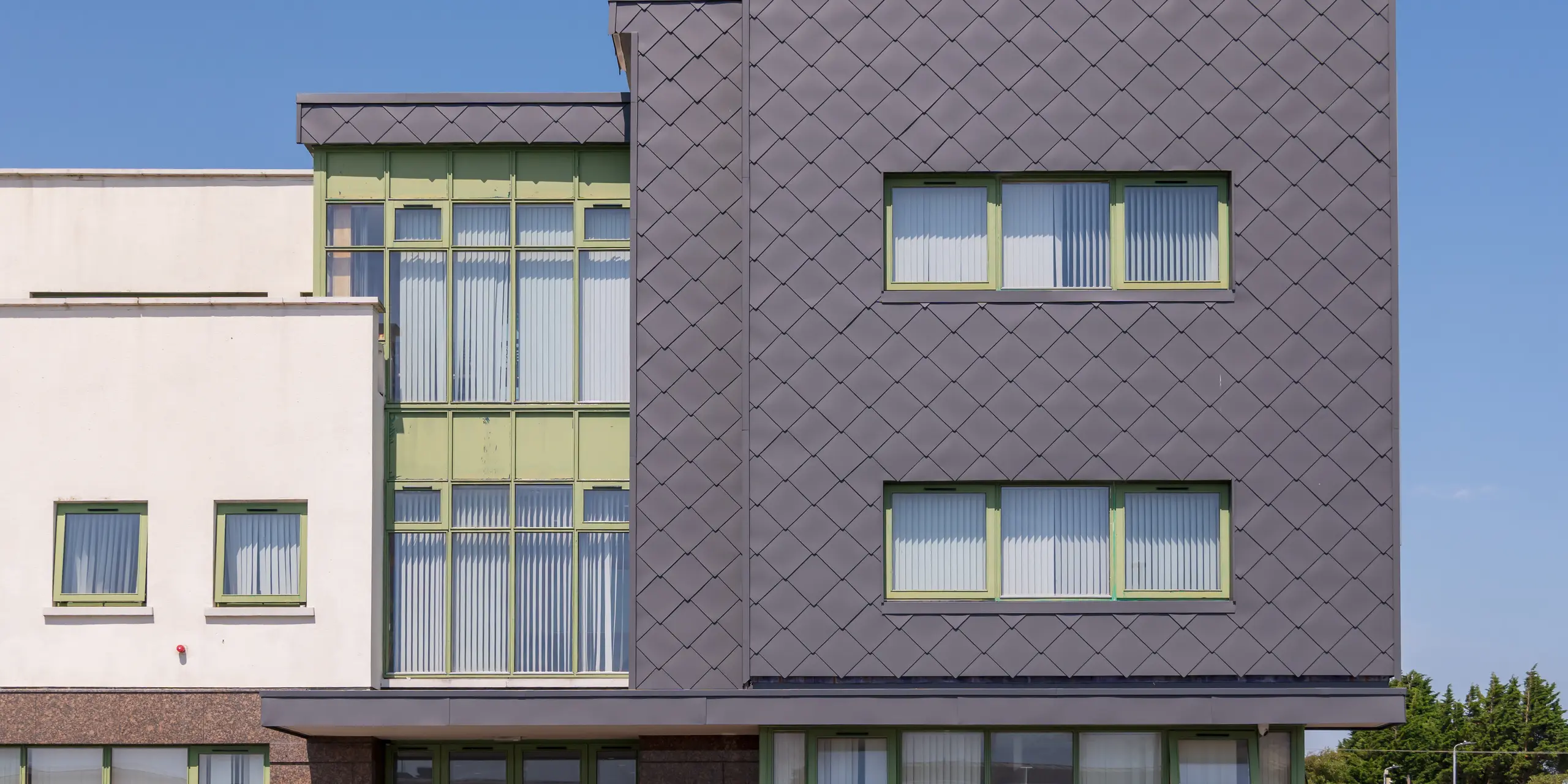 Dynamische Nahaufnahme der mit PREFA Wandrauten 44 x 44 in der Farbe P.10 Dunkelgrau verkleideten Fassade des Zentrums für psychische Gesundheit von Kindern und Jugendlichen in Wexford: Die präzise Rautenstruktur erzeugt lebendige Licht- und Schattenspiele, während die grün gerahmten Fenster Kontraste setzen. Die robuste Aluminiumfassade überzeugt durch Langlebigkeit, Wartungsarmut und hohe Witterungsbeständigkeit.