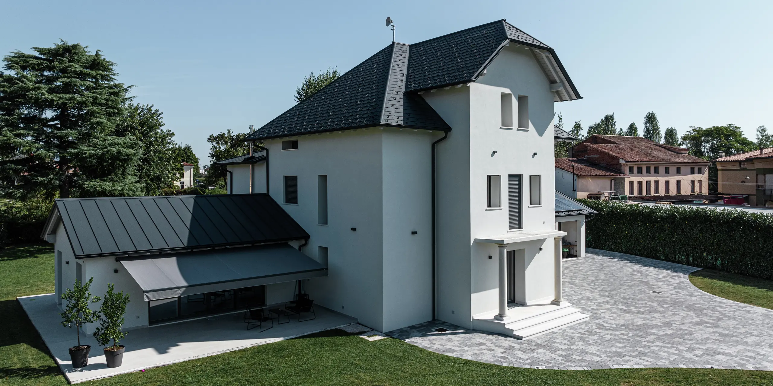 Vue latérale d'une maison familiale moderne à Marano Vicentino, en Italie, avec des bardeaux de toiture PREFA robustes en P.10 anthracite. La dépendance adjacente a été équipée d'un toit à joint debout durable. L'auvent gris clair offre de l'ombre contre le fort soleil de midi. L'architecture claire et les produits en aluminium de haute qualité garantissent un paysage de toit moderne et résistant aux intempéries.