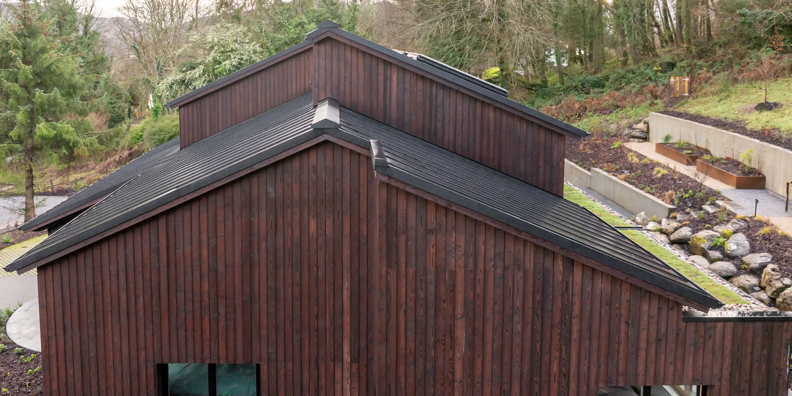 Vue latérale de la Maison Holywell à Sligo : les pignons aux formes originales guident le regard le long des lignes de toiture clairement définies, dessinées avec précision par la toiture en aluminium PREFALZ P.10 Noir. Les joints debout fins structurent les surfaces inclinées et soulignent le style architectural moderne. Un contraste saisissant se crée avec la façade en bois chaleureux. La toiture PREFA impressionne par sa durabilité, sa résistance à la corrosion et sa protection fiable même dans des conditions climatiques difficiles.