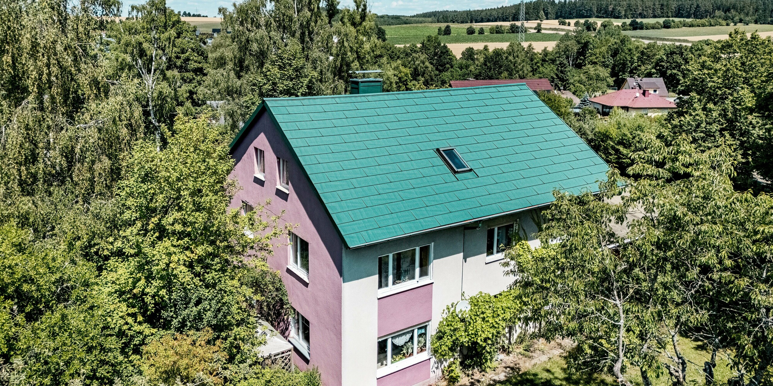 Rénovation d'une maison individuelle à Schwarzenbach-Martinlamitz avec des panneaux de toiture PREFA FX.12 couleur Vert Mousse P.10. L'image présente la toiture en aluminium modernisée, d'un vert mousse éclatant, caractérisée par ses lignes épurées et sa surface texturée. Le contraste avec la façade violette et blanche confère au bâtiment un caractère unique. Nichée dans le paysage verdoyant, la toiture apparaît à la fois moderne et en harmonie avec la nature. Durable, résistante aux intempéries et sans entretien, la toiture PREFA allie fonctionnalité et esthétique.