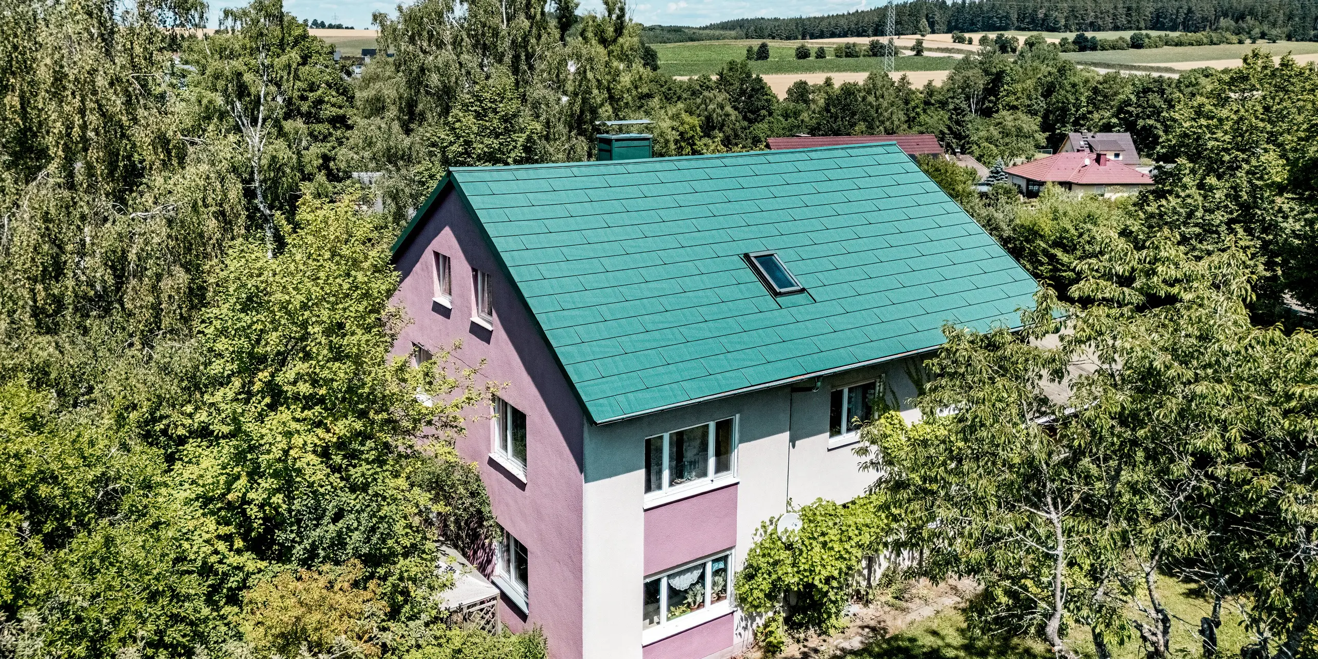 Rénovation d'une maison individuelle à Schwarzenbach-Martinlamitz avec des panneaux de toiture PREFA FX.12 couleur Vert Mousse P.10. L'image présente la toiture en aluminium modernisée, d'un vert mousse éclatant, caractérisée par ses lignes épurées et sa surface texturée. Le contraste avec la façade violette et blanche confère au bâtiment un caractère unique. Nichée dans le paysage verdoyant, la toiture apparaît à la fois moderne et en harmonie avec la nature. Durable, résistante aux intempéries et sans entretien, la toiture PREFA allie fonctionnalité et esthétique.