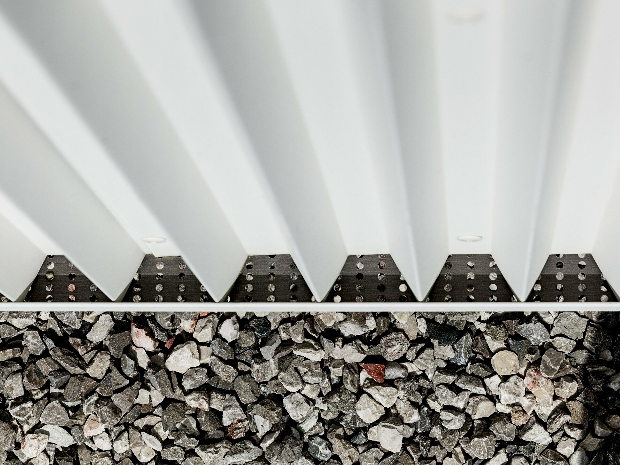 L'image présente un détail de la façade ventilée PREFALZ en aluminium blanc pur, caractérisée par des formes en zigzag pliées avec précision. Cette technique de pliage innovante garantit non seulement une structure esthétique, mais aussi une excellente ventilation et une grande résistance aux intempéries. Les éléments de façade sont assemblés à l'aide de fixations invisibles spécifiques, ce qui confère à la surface une apparence particulièrement propre et durable. La partie inférieure de l'image montre le revêtement de gravier entourant le bâtiment, assurant le drainage et la stabilisation, et soulignant ainsi la fonctionnalité de la conception de la façade. L'utilisation de PREFALZ répond aux exigences esthétiques et techniques, ce qui en fait un choix idéal pour une architecture moderne et durable.