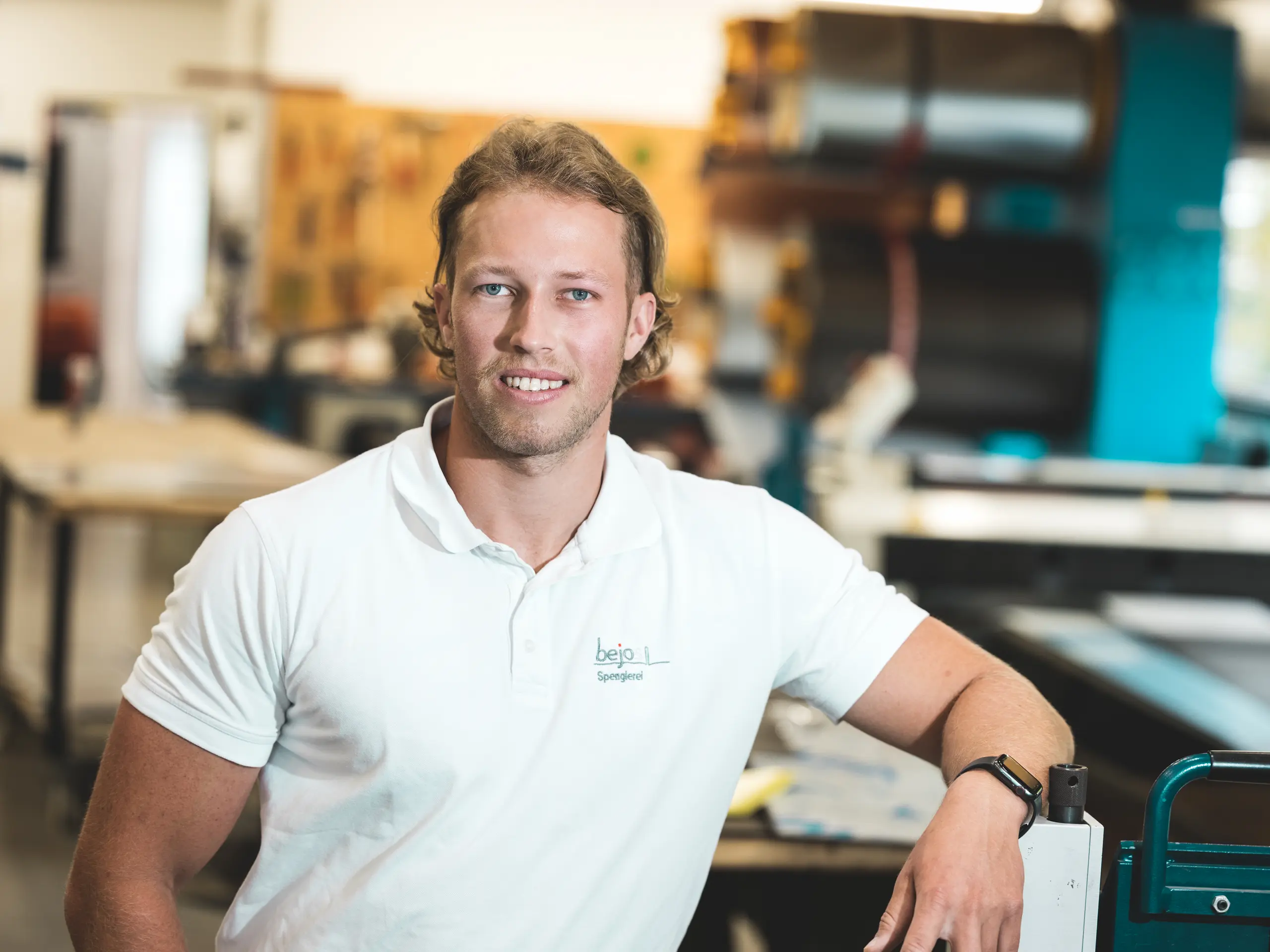 Portrait de Jakob Berchtold, maître couvreur-zingueur de l’entreprise vorarlbergeoise Bejos, dans son atelier. L’artisan s’appuie nonchalamment contre une machine et regarde l’objectif en souriant. Pour la maison Immi, il a réalisé la toiture solaire PREFA intégrée, composée de tuiles solaires et de panneaux de toiture R.16 en P.10 noir : une solution robuste, facile à poser et à l’esthétique harmonieuse.